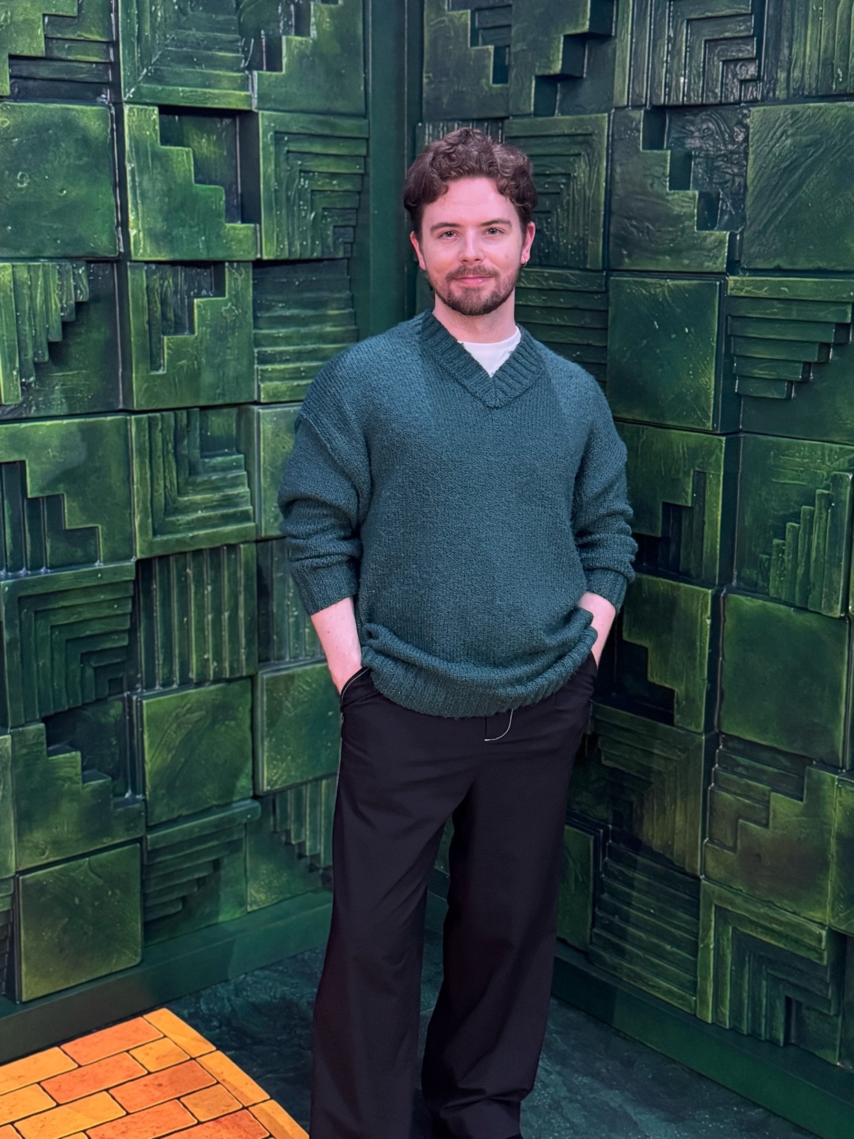 A man poses in front of a green geometric wall. He wears a green sweater and black pants.