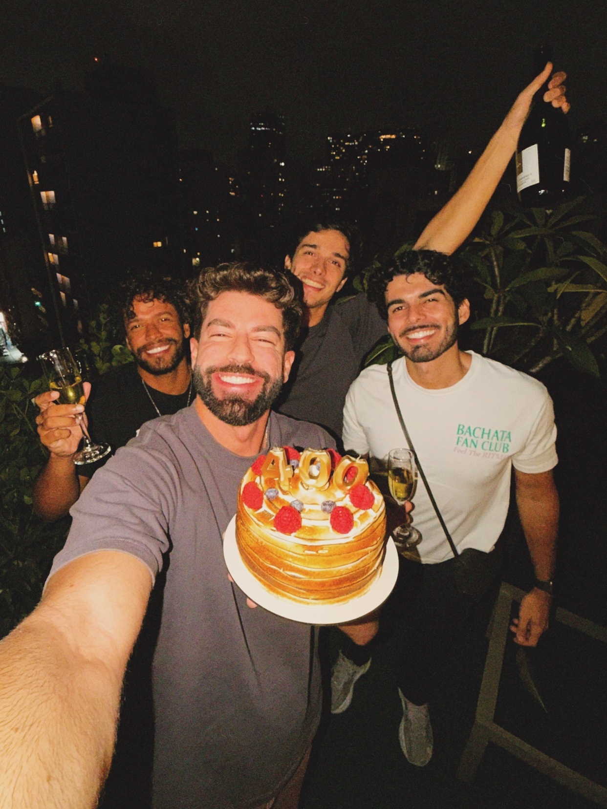 Four smiling men pose for a selfie at night, holding a birthday cake and champagne. One man in the foreground holds a cake decorated with raspberries and the number '400', while another raises a bottle of champagne in the background.