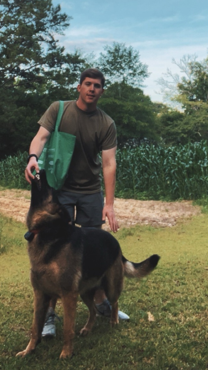 A man in a green t-shirt and shorts stands next to his German Shepherd dog in a grassy, outdoor setting.