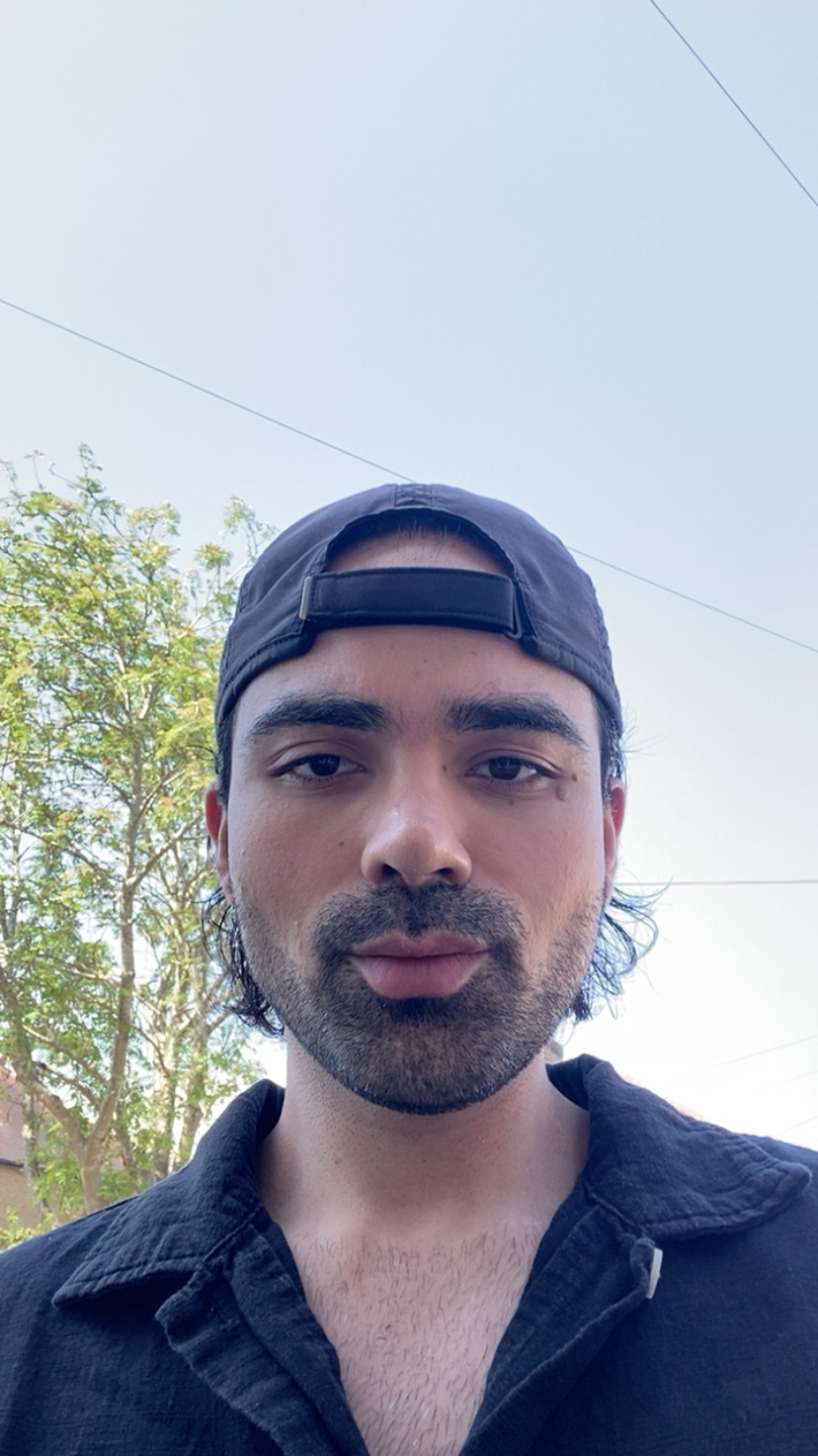A man wearing a black baseball cap and a black collared shirt poses for a selfie outdoors. He has stubble and a beard, with dark hair visible from under his cap.