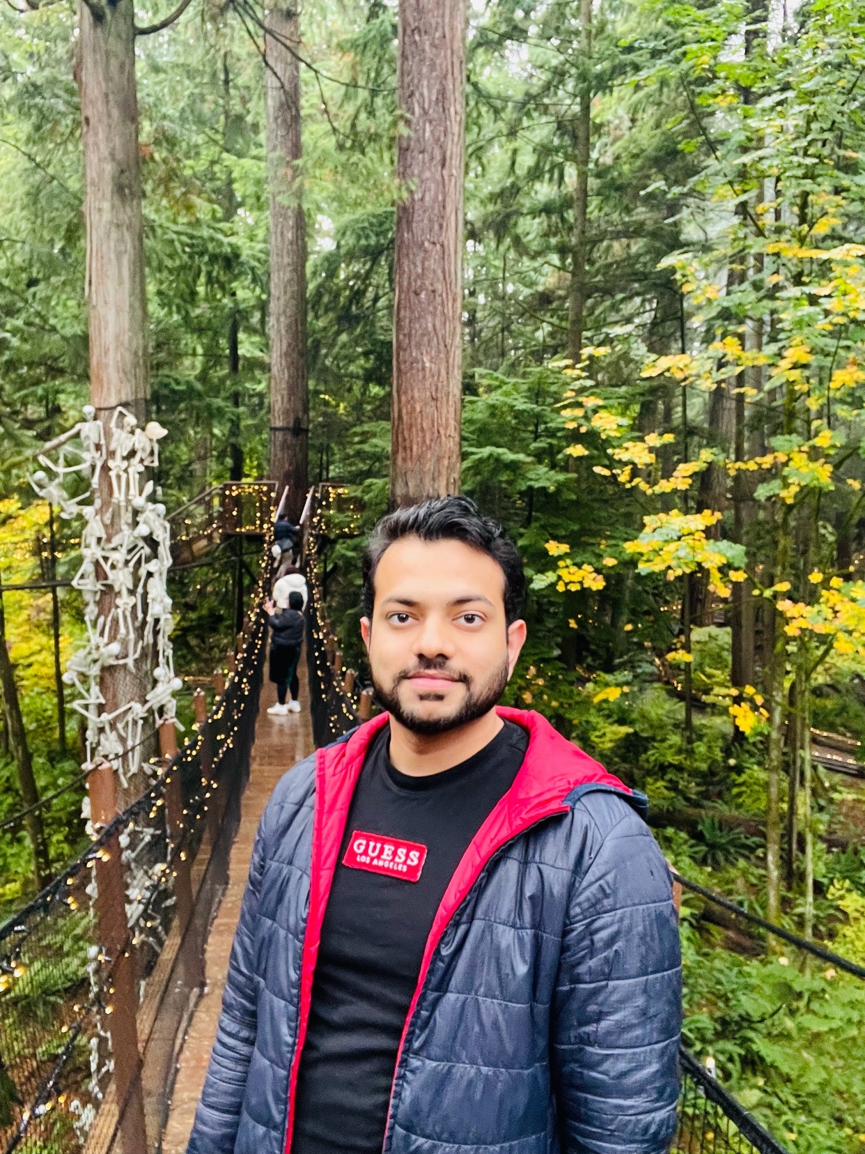 A man poses on a bridge in a lush forest. He is smiling and wearing a jacket.