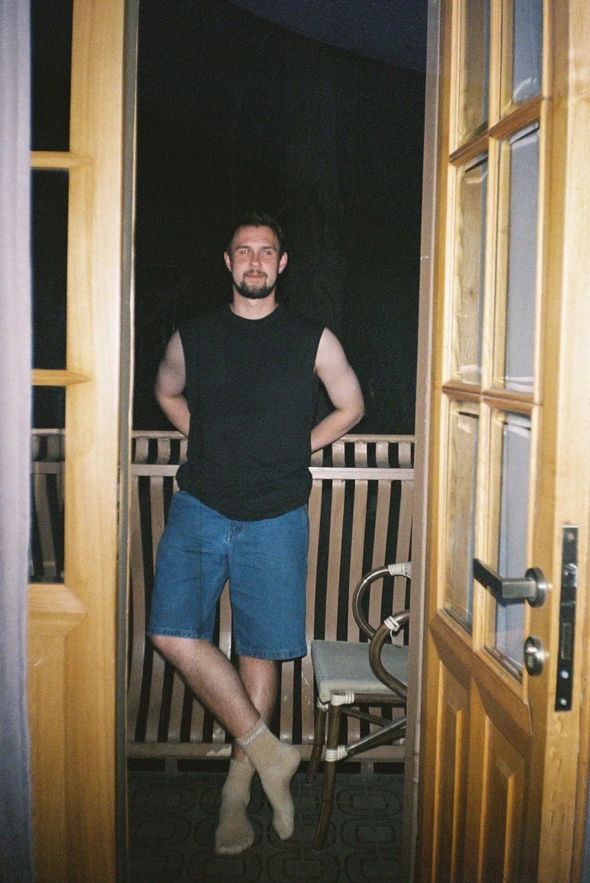 A man stands on a balcony, leaning against a railing. He is wearing shorts and a sleeveless shirt.