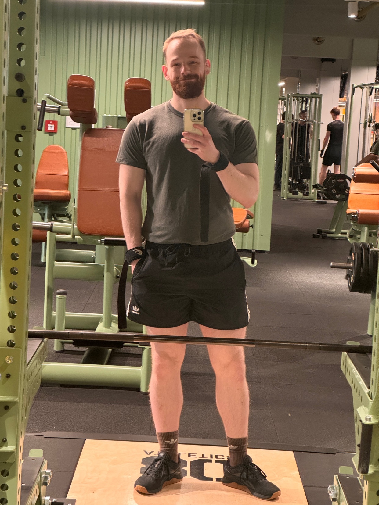 A man takes a mirror selfie at the gym, showcasing his physique. He's wearing workout clothes and appears to be ready for a session.
