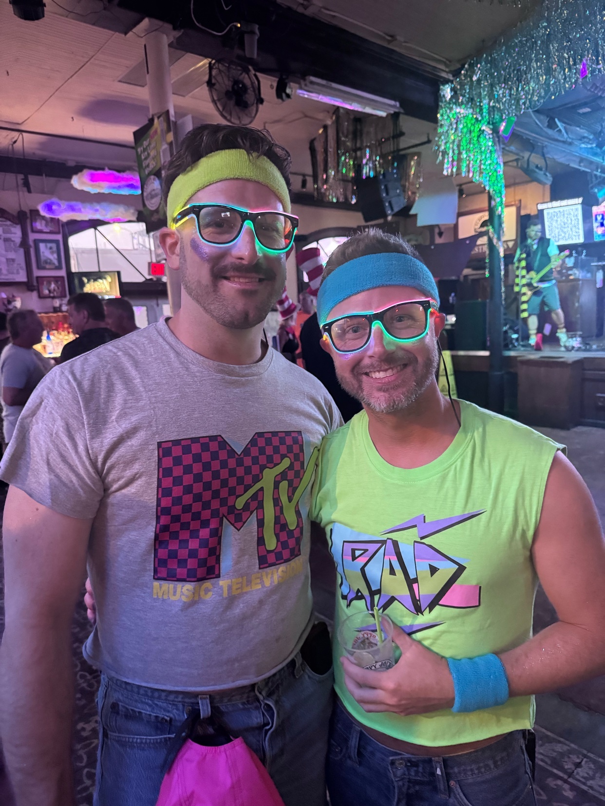 Two men pose for a photo at a party, dressed in 80s-themed costumes. They wear neon accessories and shirts with retro logos.