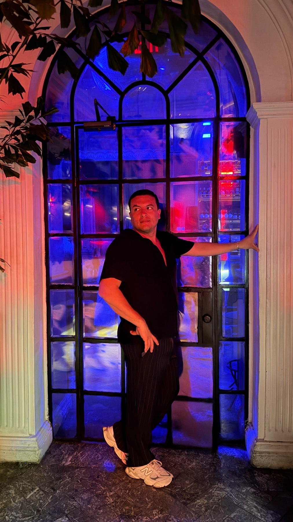 A man stands in a doorway illuminated by vibrant blue and red lights. He is wearing a black collared shirt and striped pants, with his hand resting on the doorframe.