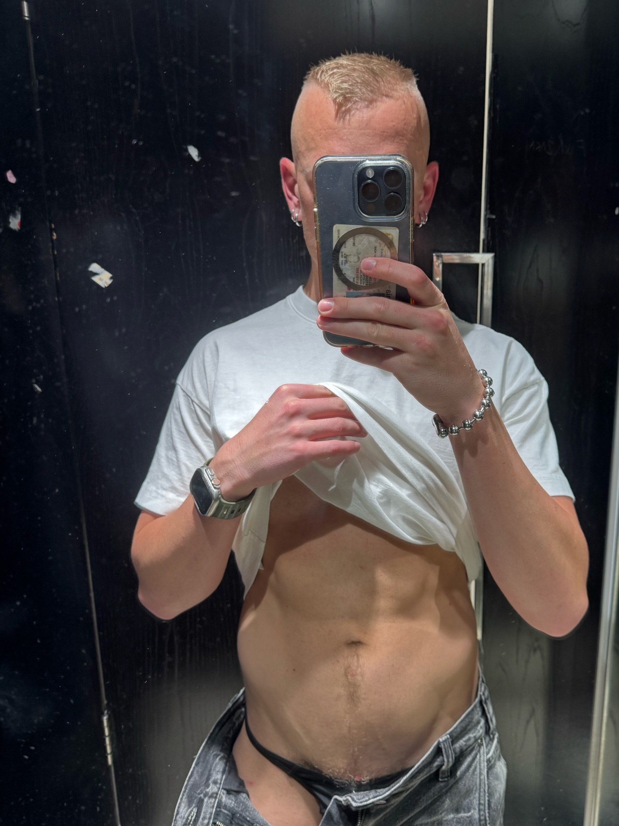 A man with a mohawk and blond hair takes a selfie in a mirror, lifting his t-shirt to reveal his abs and underwear.