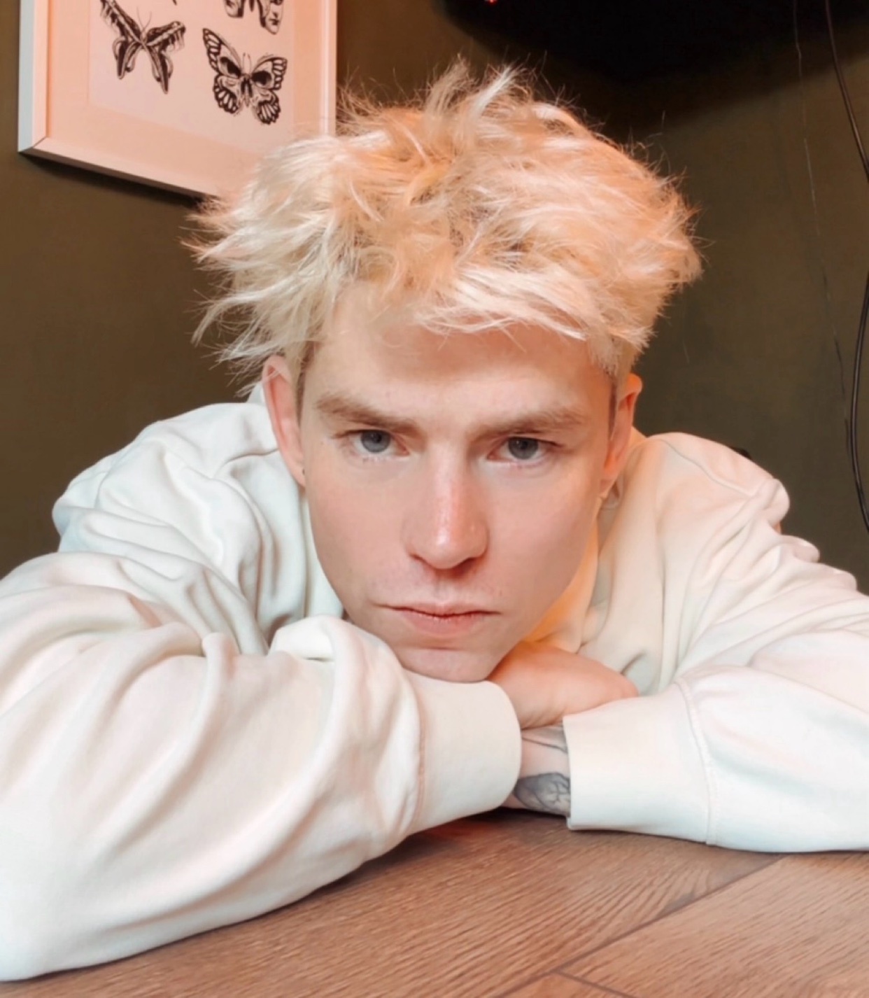 A close-up shot of a young man with messy, blonde hair and a serious expression. He is wearing a cream-colored hoodie and resting his chin on his hands.