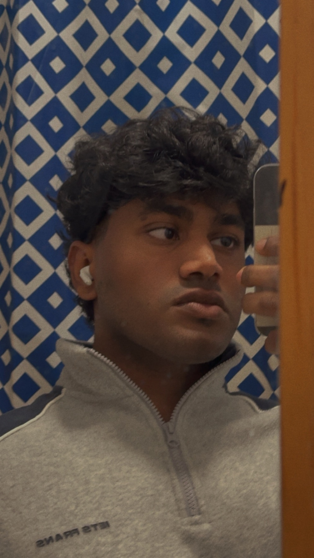 A man takes a mirror selfie. He has dark curly hair and is wearing a grey sweater.