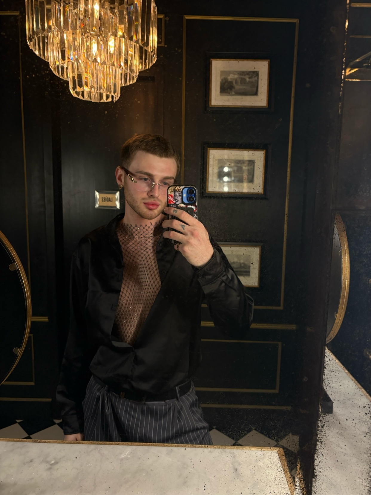 A man takes a mirror selfie in a stylish outfit. He is shirtless under a black satin shirt.
