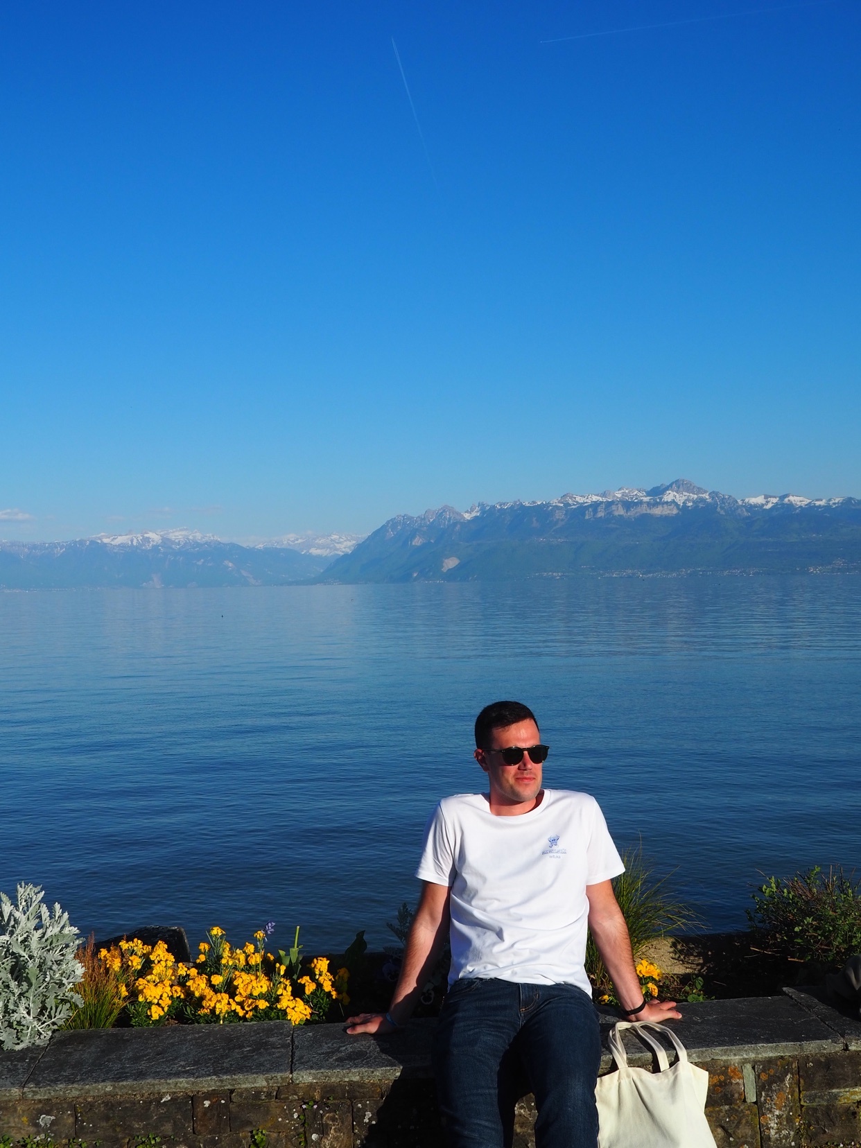 A man wearing sunglasses and a white t-shirt sits on a stone wall overlooking a large body of water with mountains in the distance under a clear blue sky.