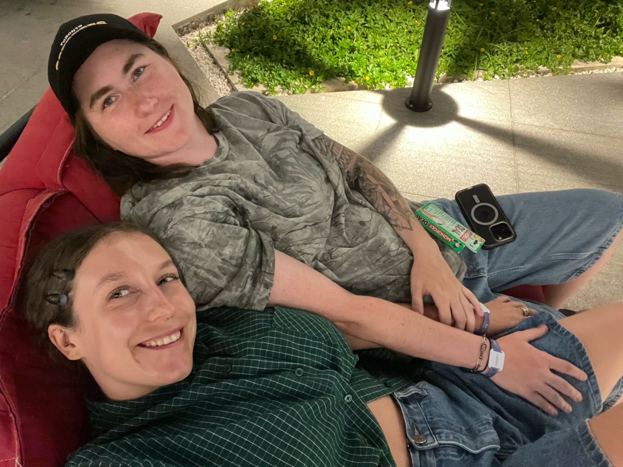 A close-up, low-angle shot shows two smiling women resting together on a red couch. The woman in the foreground wears a green plaid shirt, while the woman behind her has a grey t-shirt and a black baseball cap.