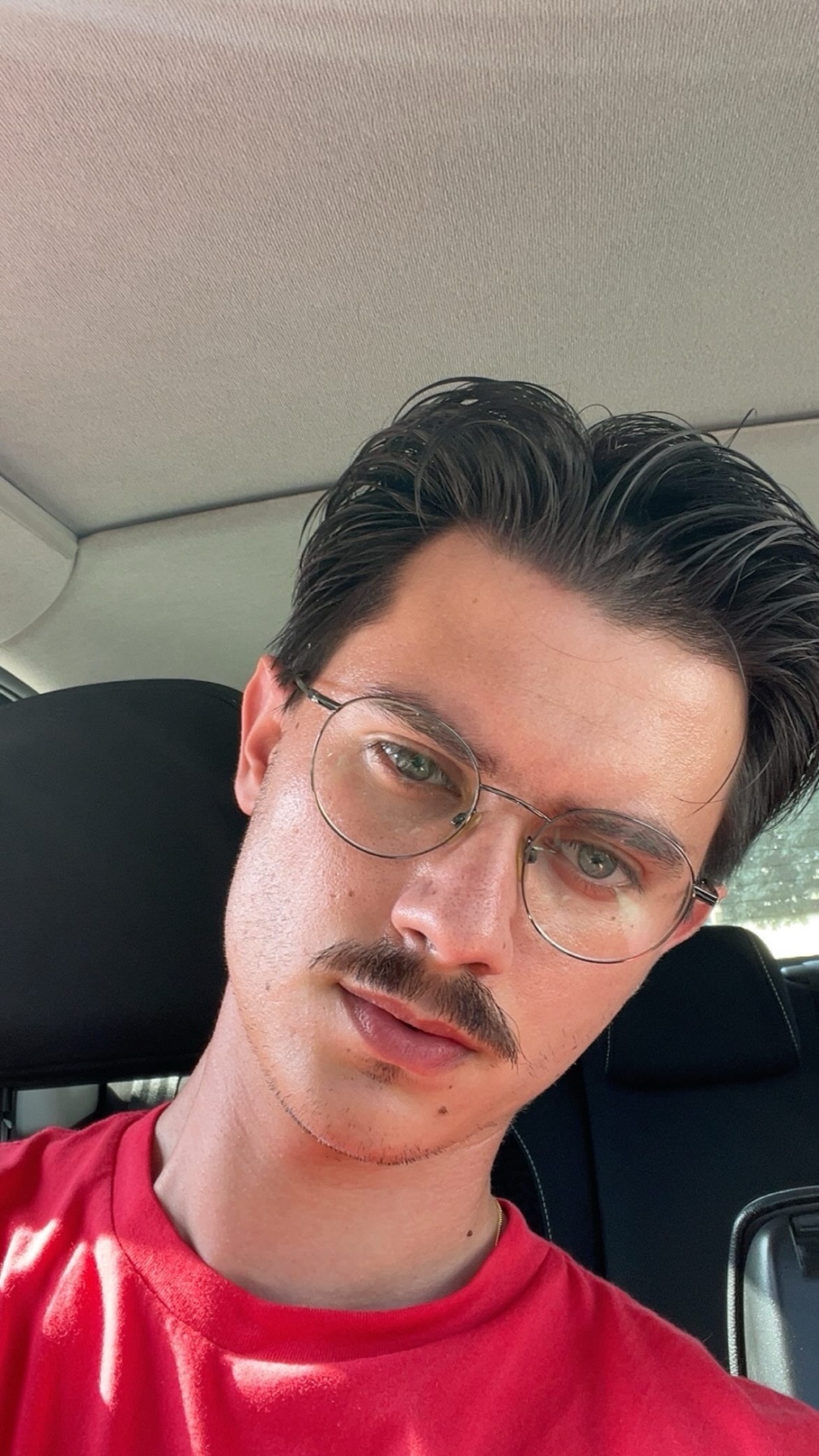 A man with a mustache and glasses poses for a selfie in a car, wearing a red shirt.