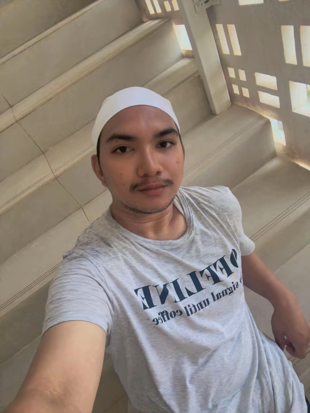 A man with stubble and a moustache takes a selfie while wearing a white kufi and a grey t-shirt. He is standing on a staircase.