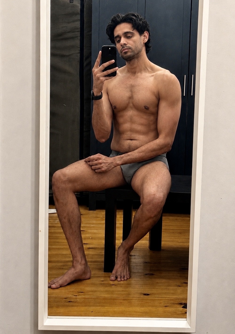 A man with dark, wavy hair takes a mirror selfie while wearing grey underwear. He is seated and looking at his phone, with his torso bare and muscular.