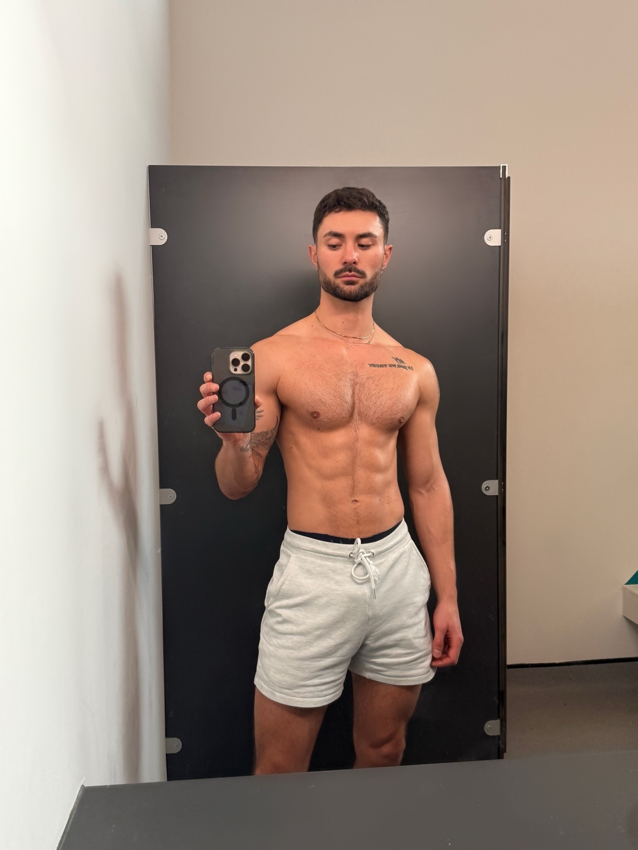 A man takes a mirror selfie, showcasing his muscular physique. He is shirtless, wearing shorts, and has a tattoo on his chest.