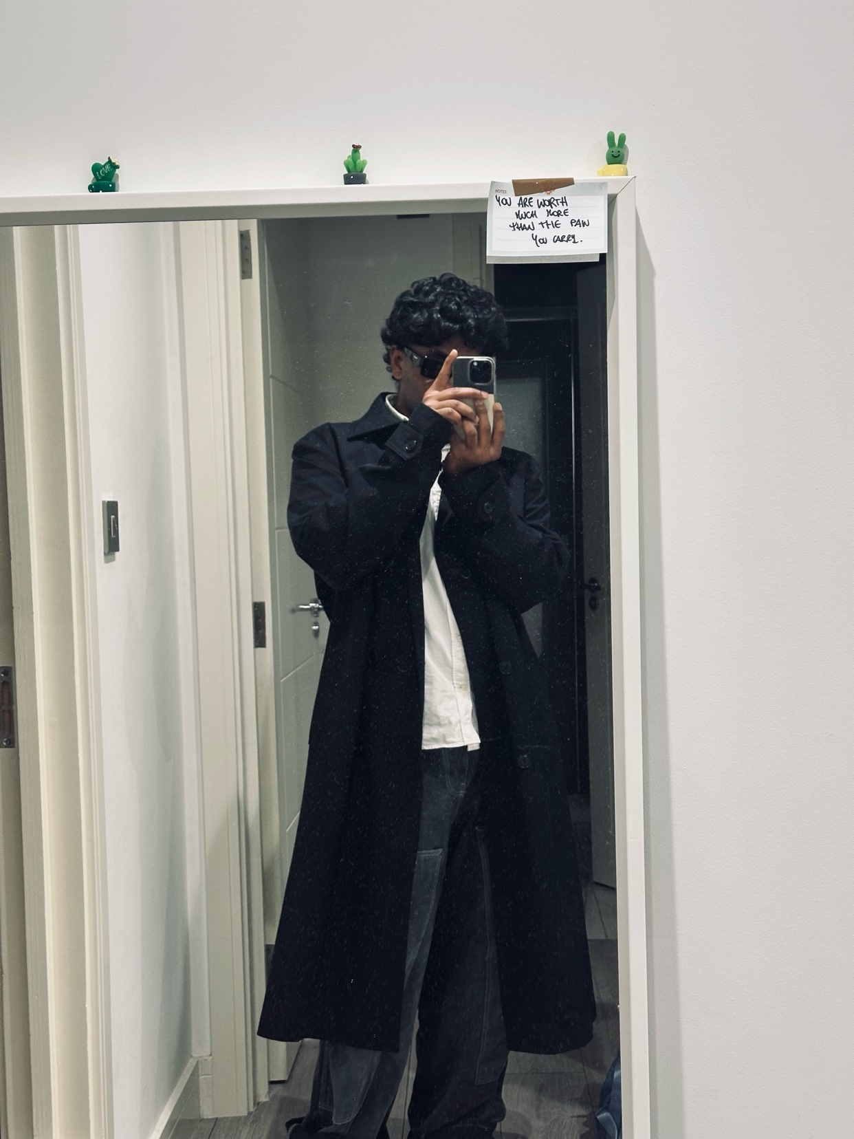 A man takes a mirror selfie, showcasing his outfit. He's wearing a long black coat and glasses.