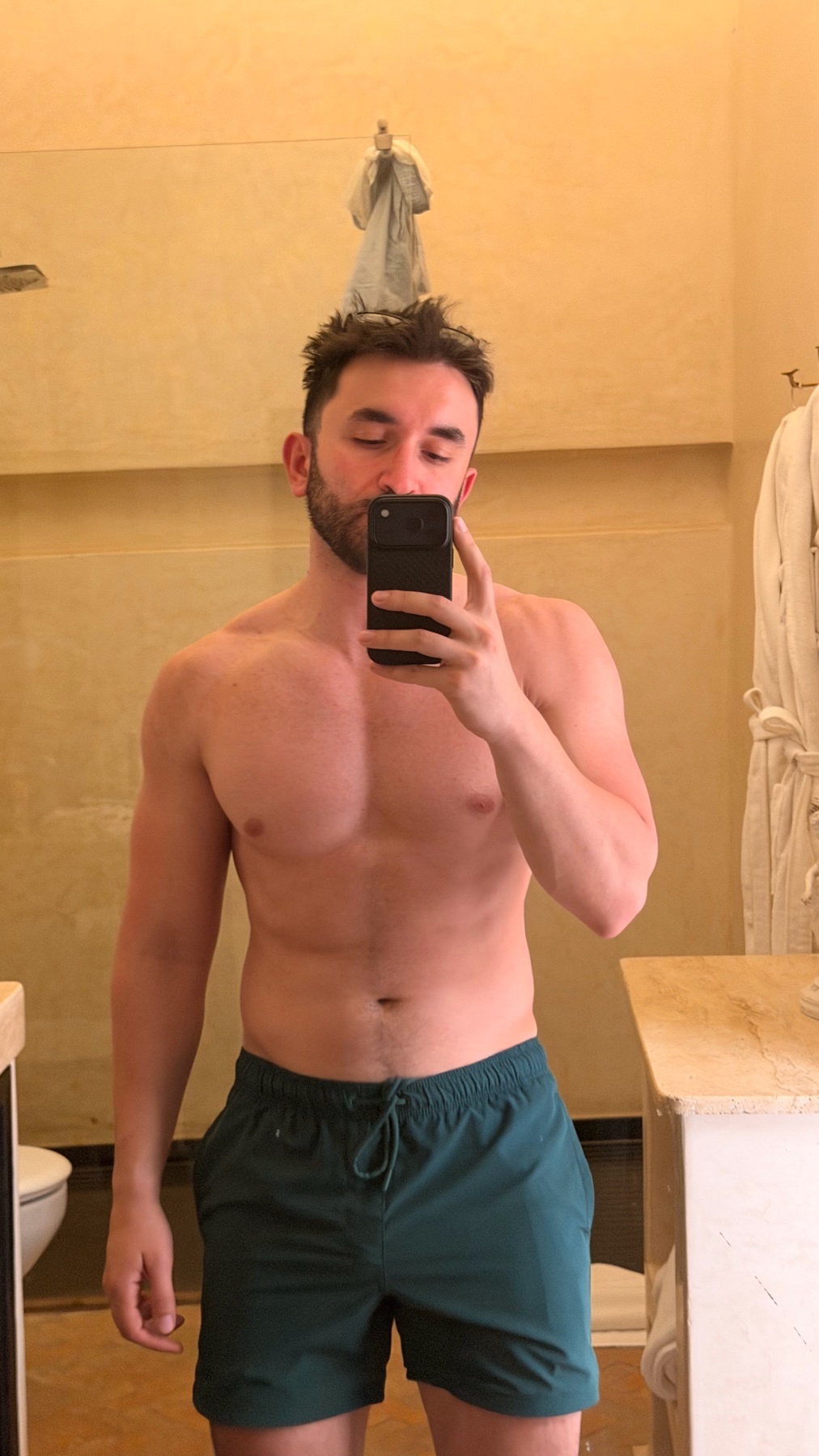 A man with a beard and dark hair takes a mirror selfie in a bathroom. He is wearing dark green swim trunks and appears muscular.