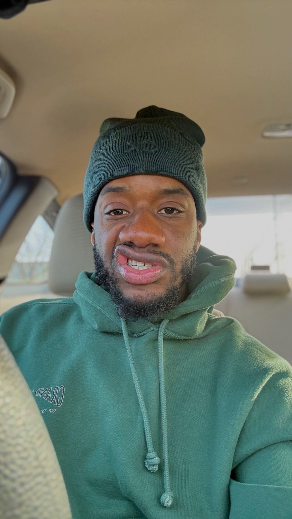 A man makes a funny face while taking a selfie in a car. He is wearing a green beanie and hoodie.