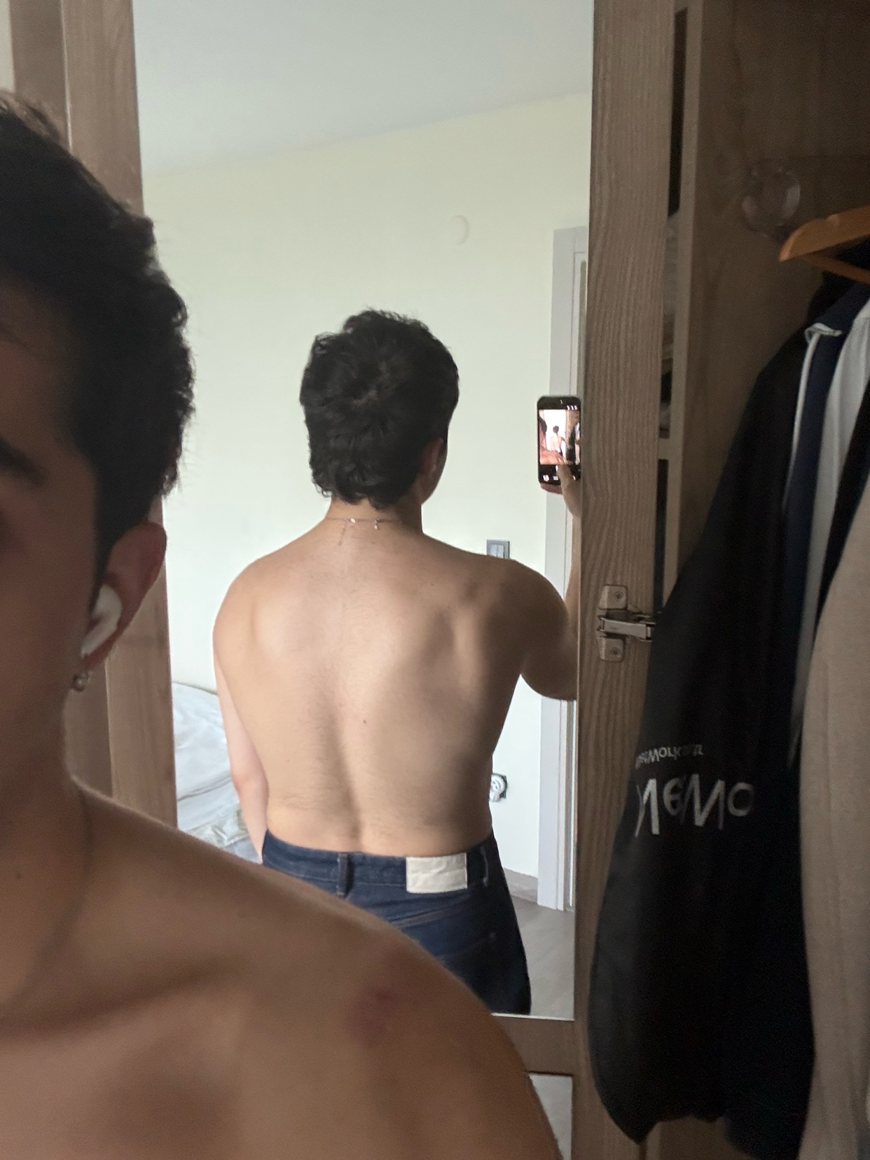 A man takes a mirror selfie, showcasing his back and physique. He is shirtless, wearing jeans, and appears to be in a gym or workout setting.