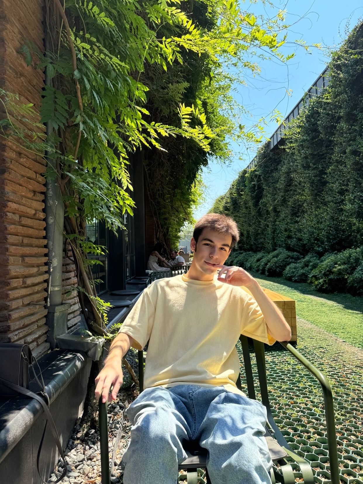 A young man poses outdoors, seated in a chair. The photo is taken in daylight, with greenery and architecture in the background.