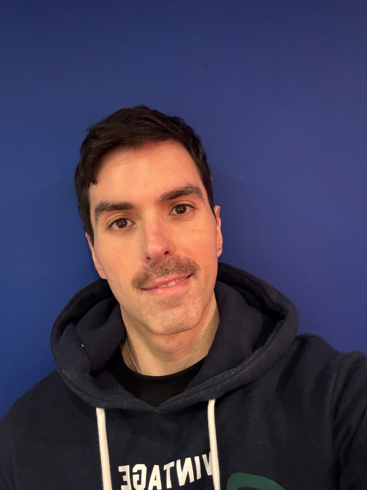 A man with dark hair and a mustache poses for a selfie. He is wearing a hoodie against a blue background.