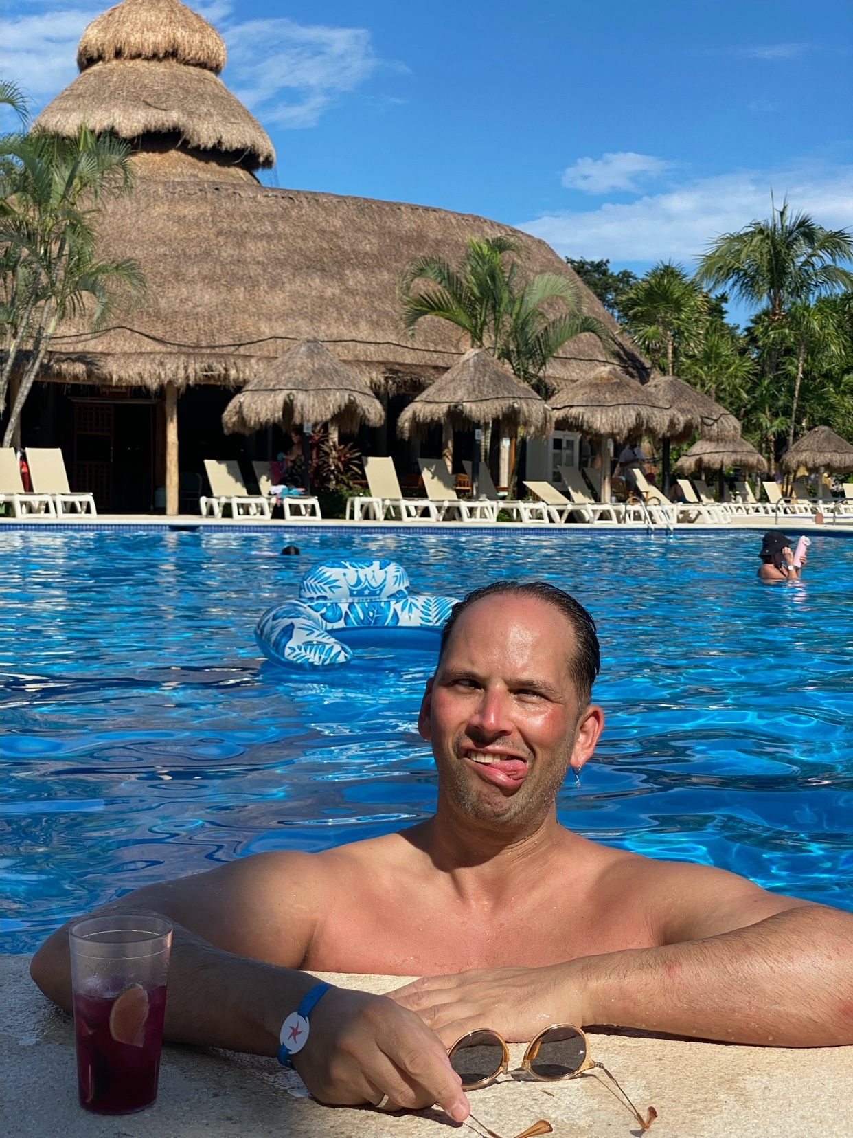 A man is in a pool, sticking his tongue out. He is shirtless and appears to be on vacation.