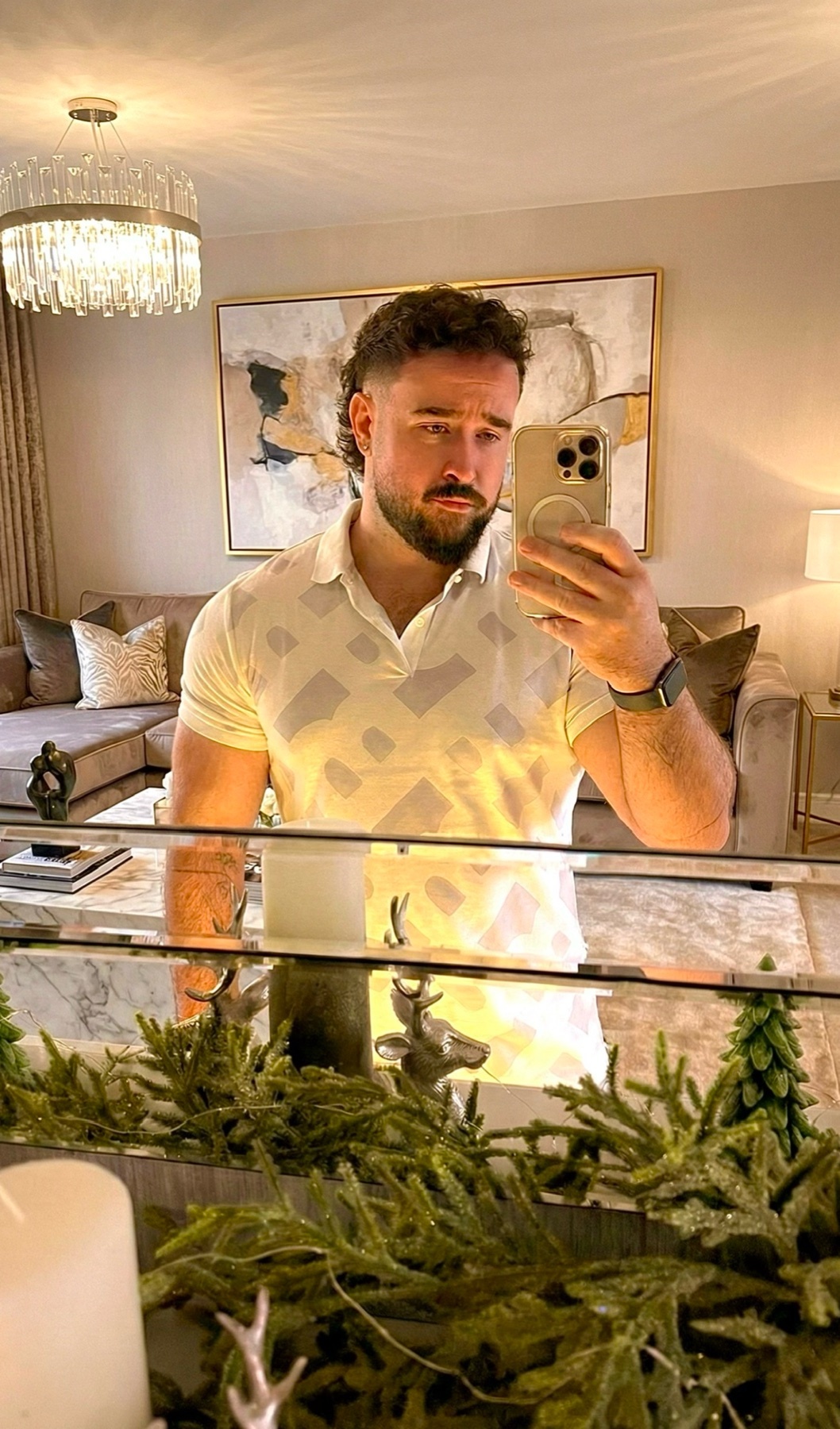 A man takes a mirror selfie in a stylish home. He is wearing a polo shirt and has a beard.