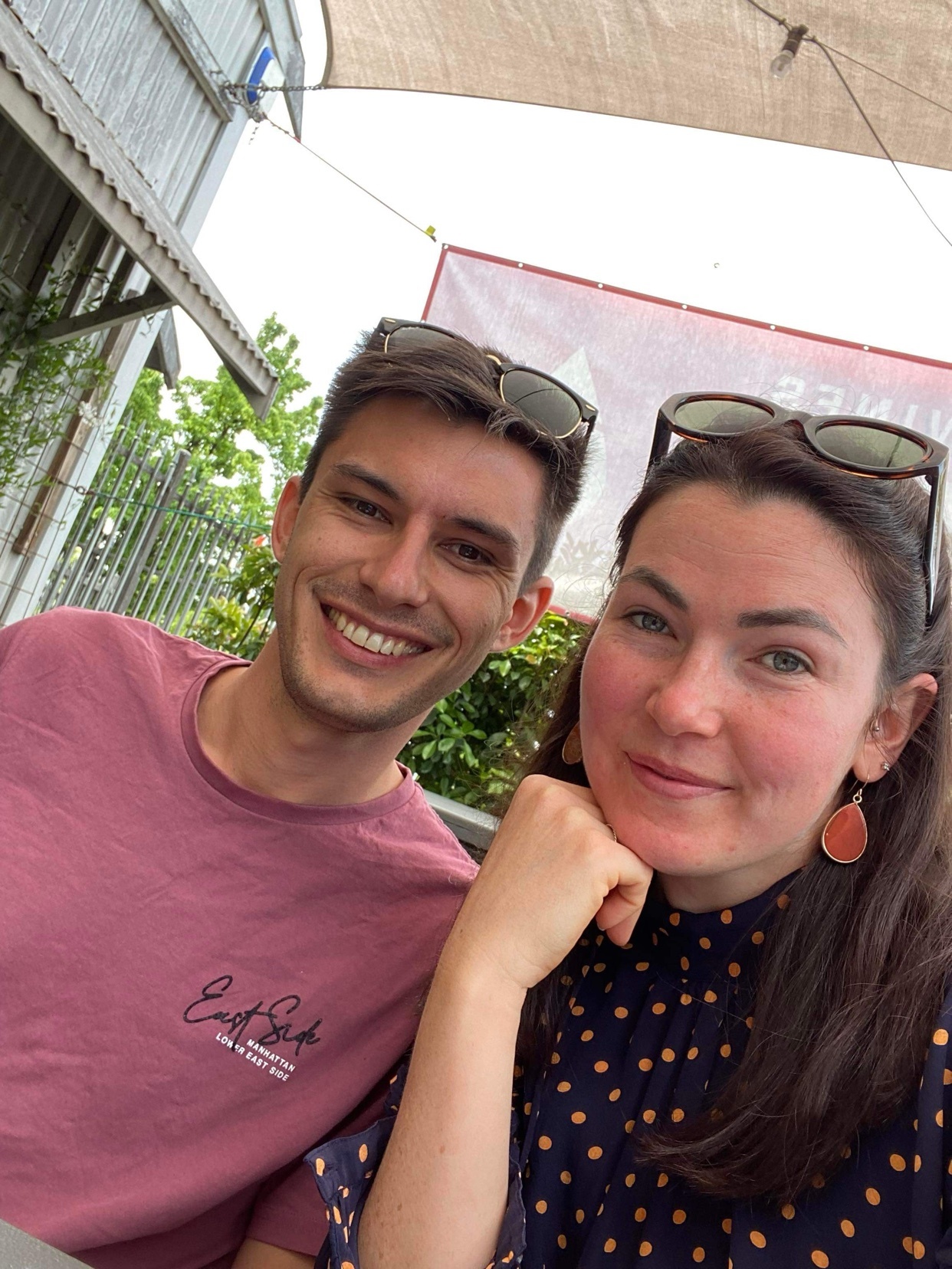 A smiling couple poses for a selfie outdoors. The man on the left wears a maroon t-shirt, and the woman on the right wears a navy polka dot dress.