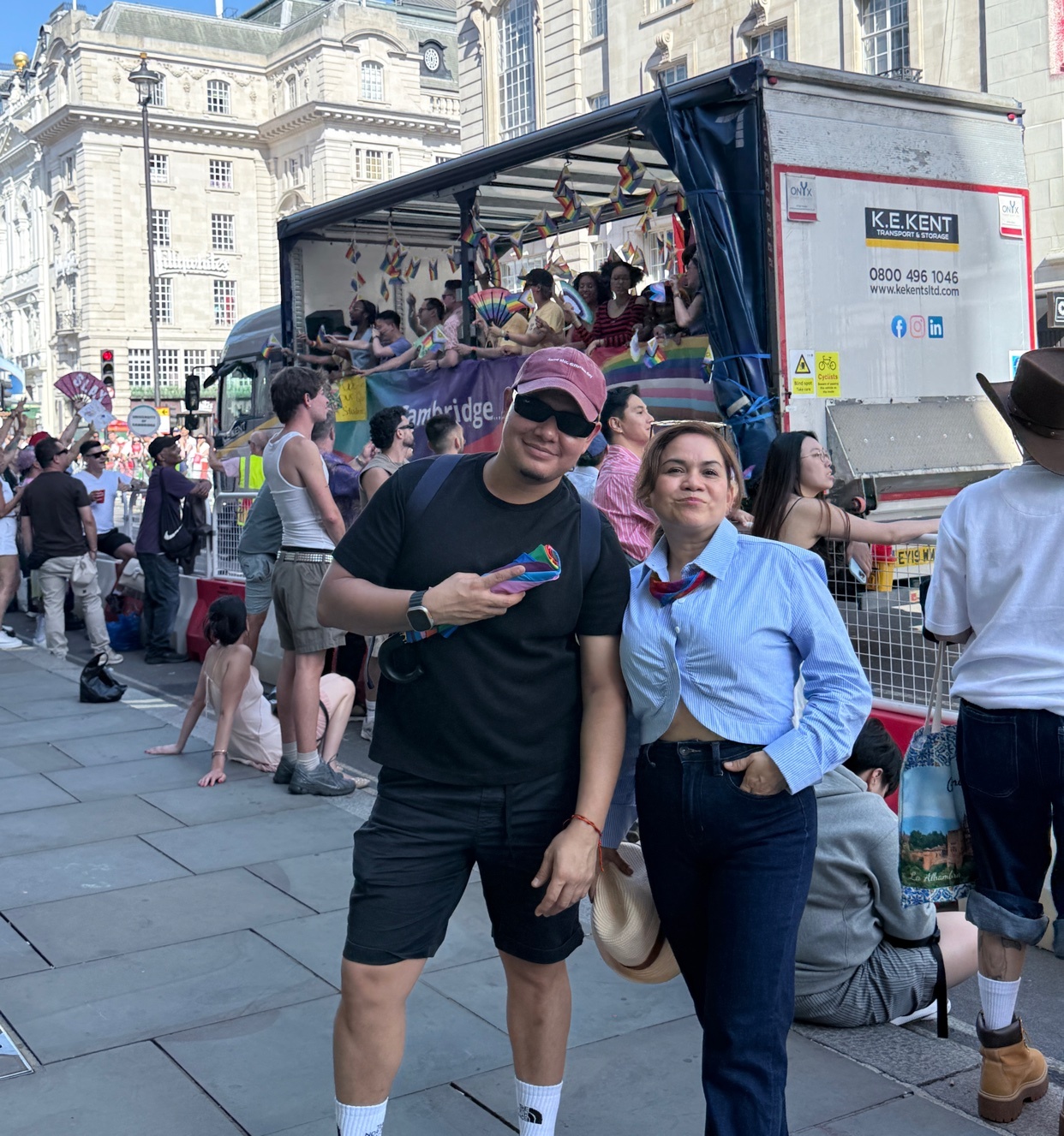 A man and a woman pose for a photo in front of a truck decorated with pride flags during a parade. The man wears a baseball cap and sunglasses, while the woman has her hair pulled back and wears a blue collared shirt.