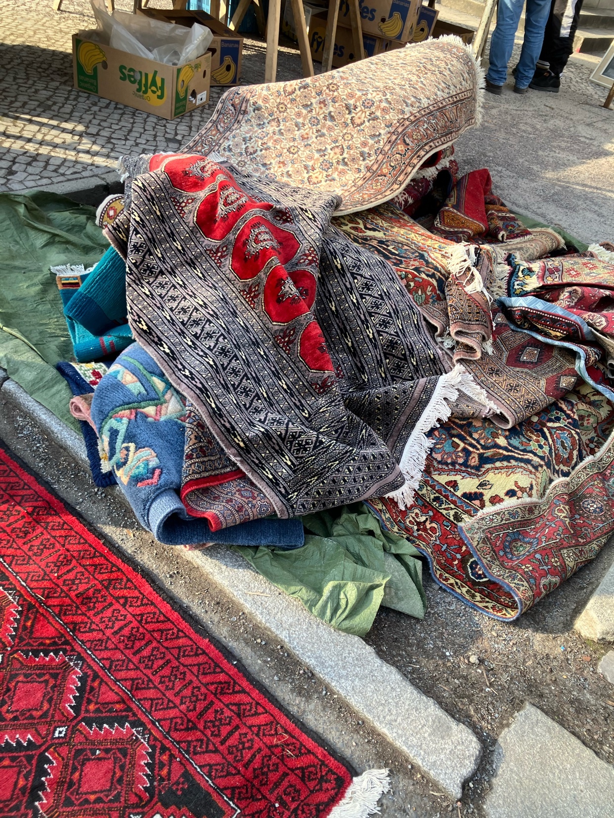 A pile of colorful, patterned rugs and carpets are displayed on the ground, some folded and others spread out. The scene appears to be at an outdoor market, with a box labeled 'Fiffes' visible in the background.