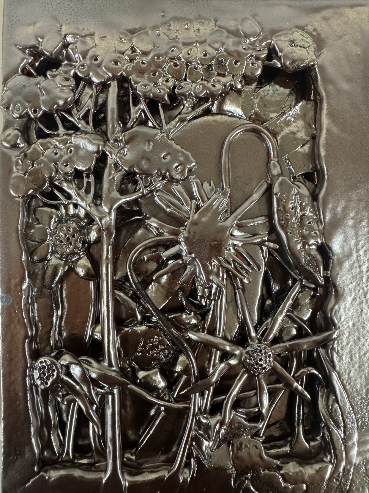 A close-up of a metal sculpture depicting a stylized floral and botanical scene. The artwork has a textured surface and a reflective, silver finish.
