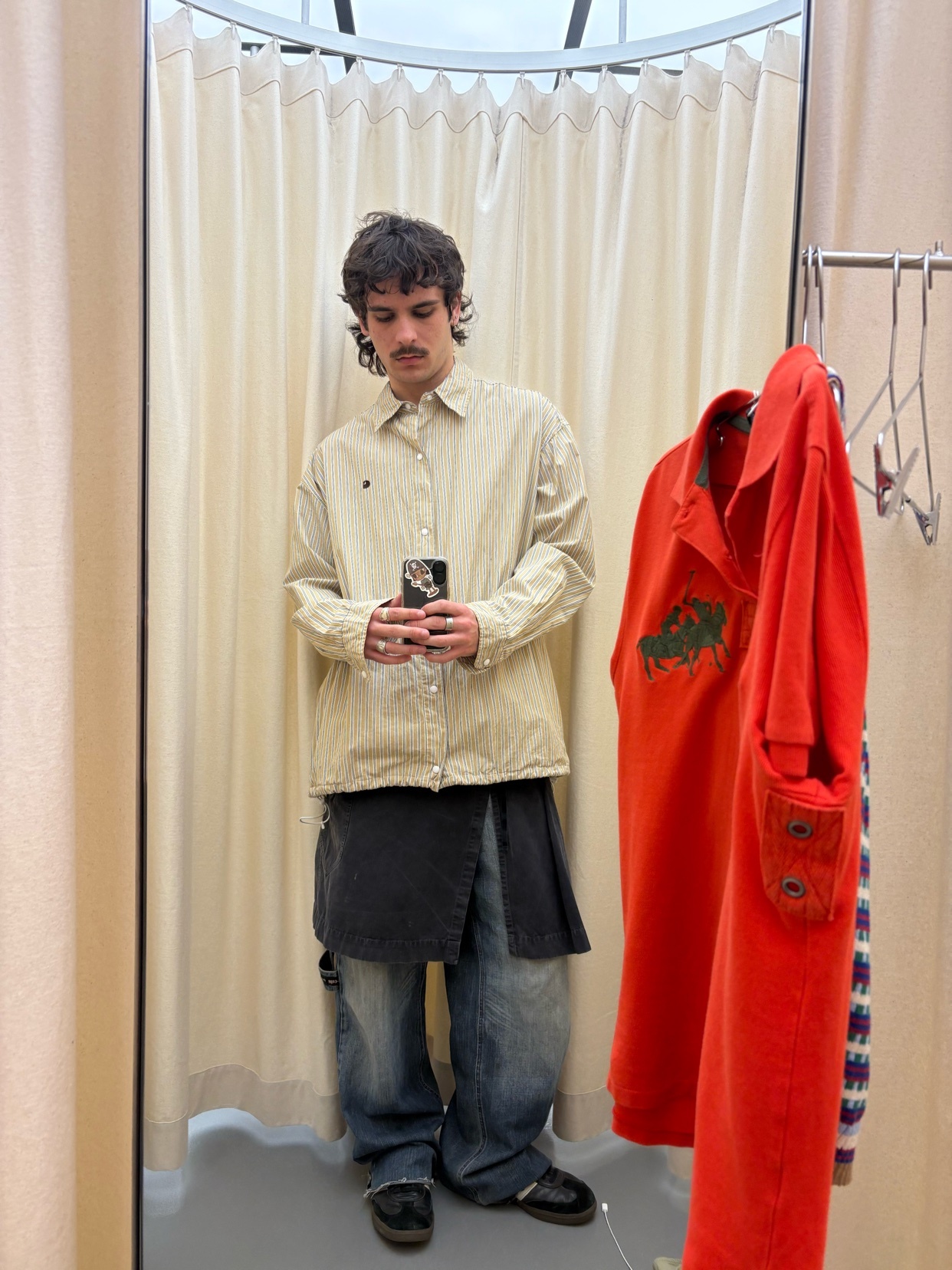 A man takes a mirror selfie in a dressing room, showcasing his outfit. He wears a light-colored shirt, baggy jeans, and stylish shoes.