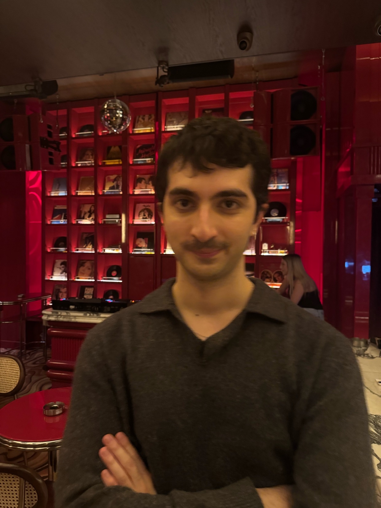 A man poses for a portrait in a restaurant. He is wearing a sweater and has dark, curly hair.
