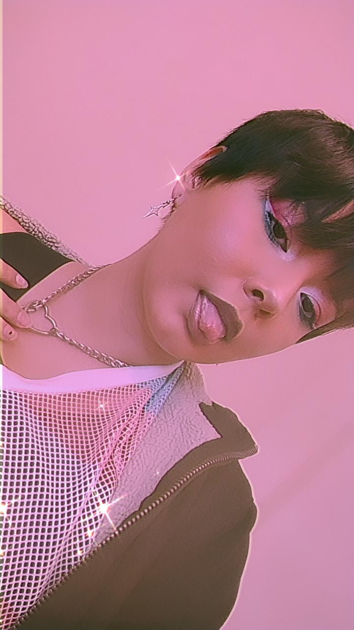 A close-up, low-angle shot of a woman with short, dark hair and bold eye makeup. She is sticking her tongue out slightly, and the image has a pink, glittery aesthetic.