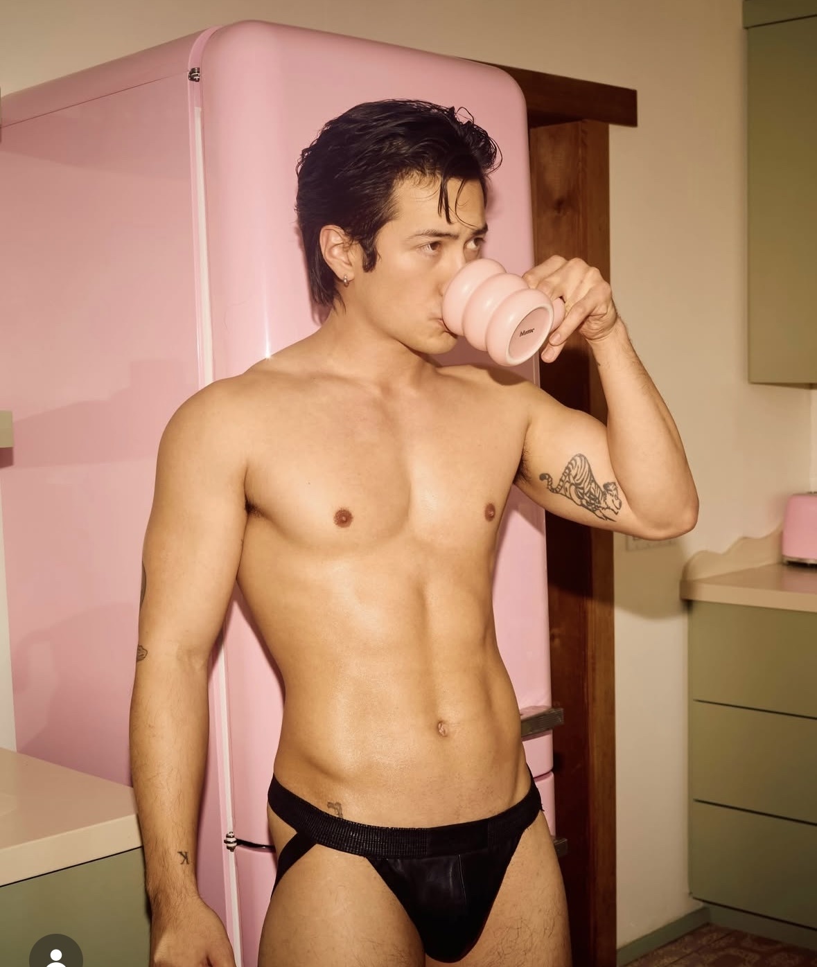 A man is shirtless, wearing underwear, and drinking from a mug in a kitchen. The photo has a modern aesthetic.