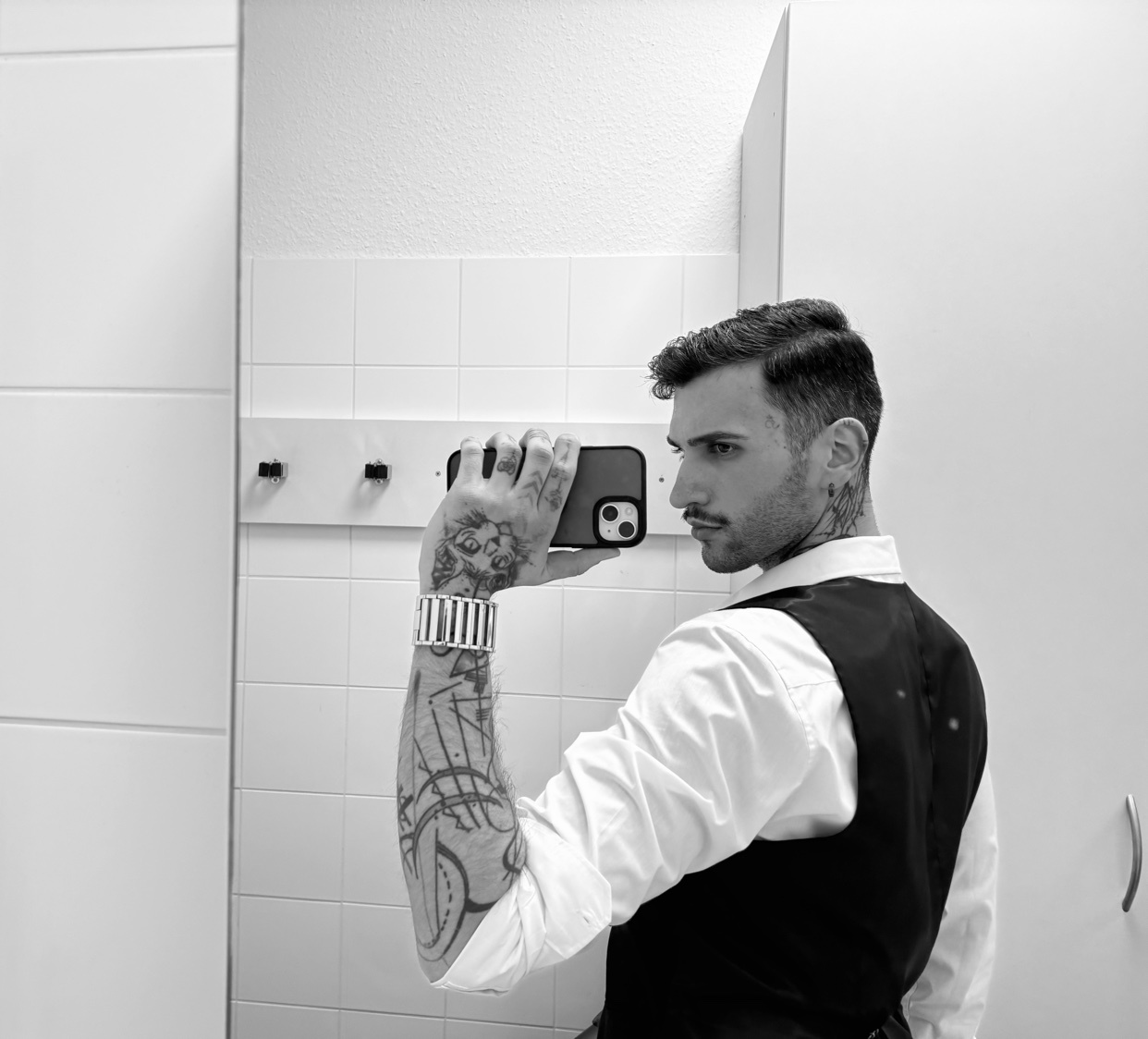 A man takes a mirror selfie, showcasing his tattoos and stylish attire. The black and white photo highlights his formal wear and modern aesthetic.