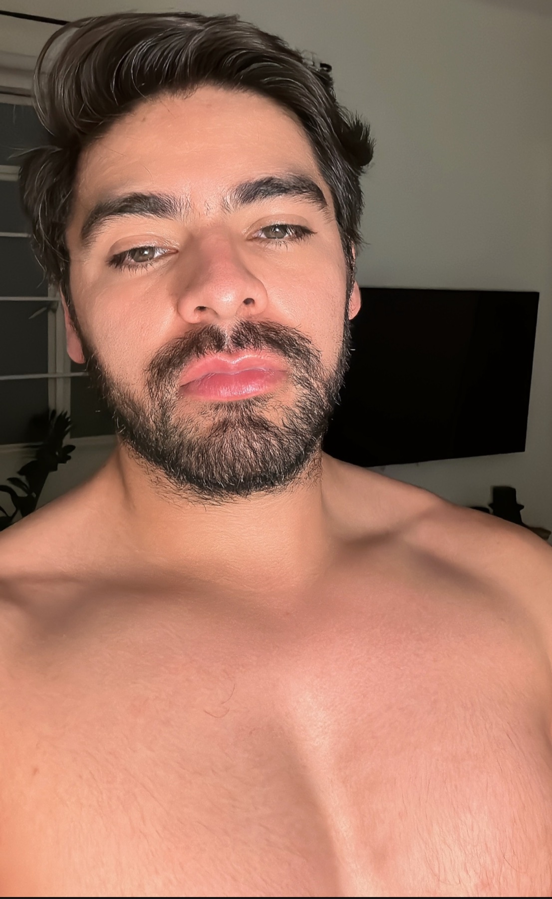 A close-up, chest-up selfie of a man with dark hair and stubble. He is shirtless and looking directly at the camera with a pouty expression.