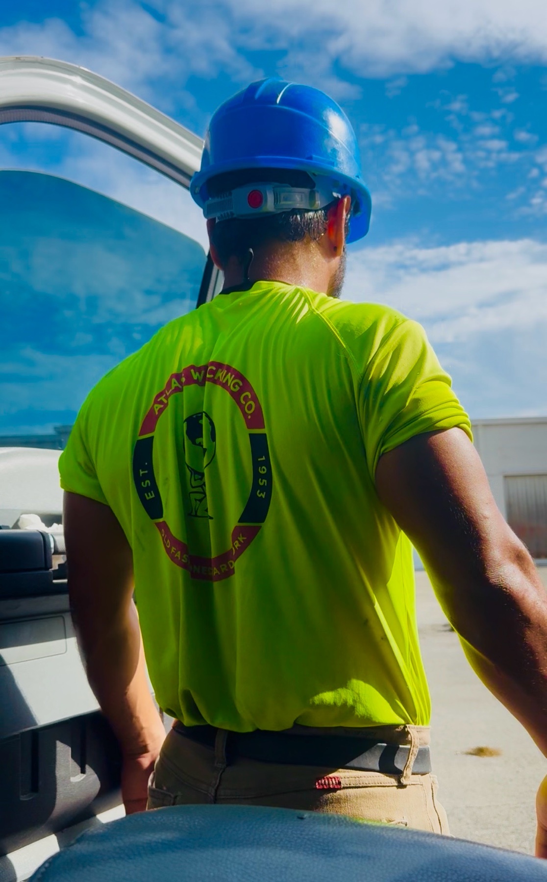 A man wearing a blue hard hat and a bright yellow t-shirt with a company logo on the back stands next to a vehicle. The image is taken from behind, focusing on his muscular build and work attire against a clear blue sky.