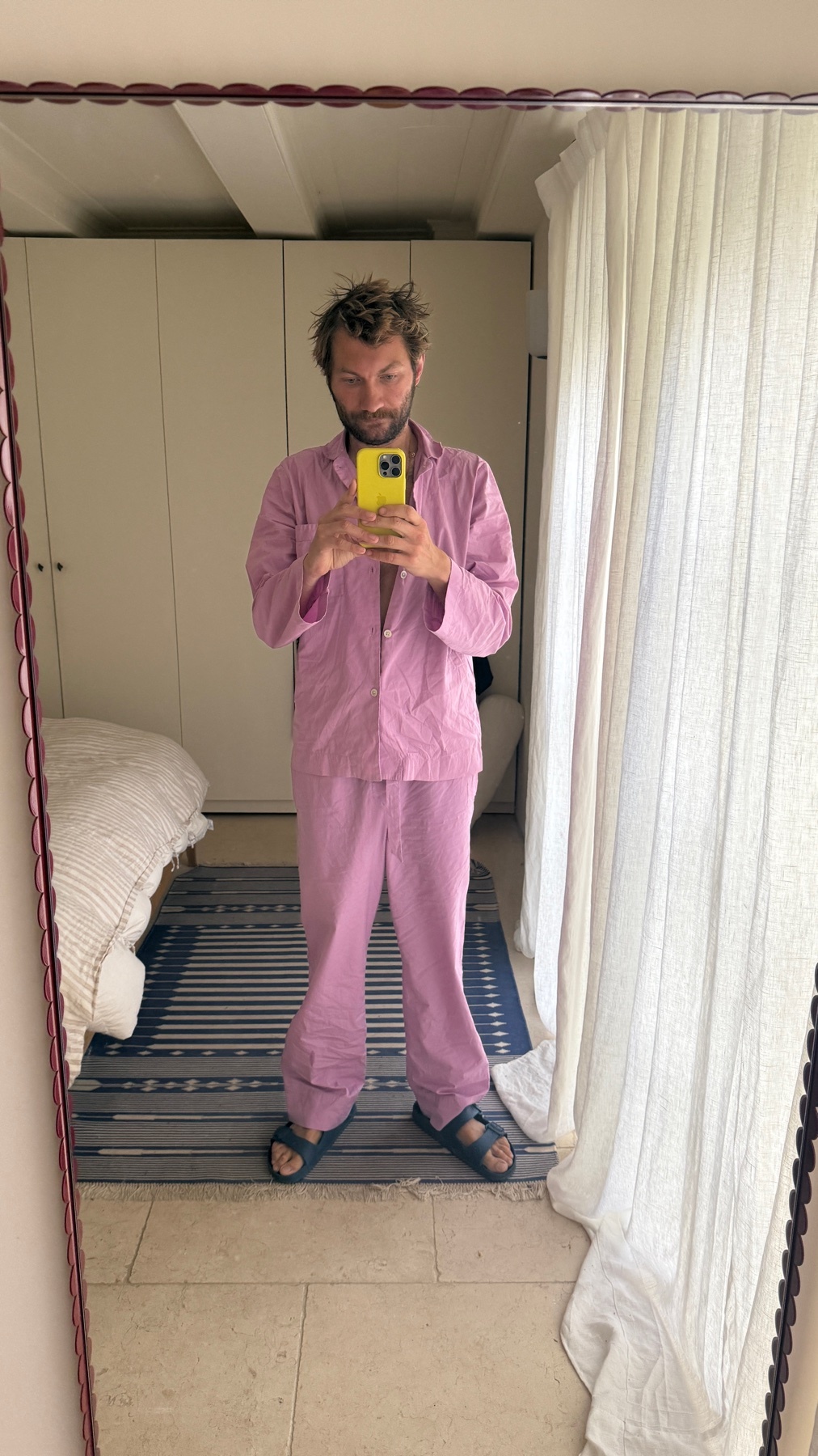 A man takes a mirror selfie in pink pajamas. He has curly hair and a beard.