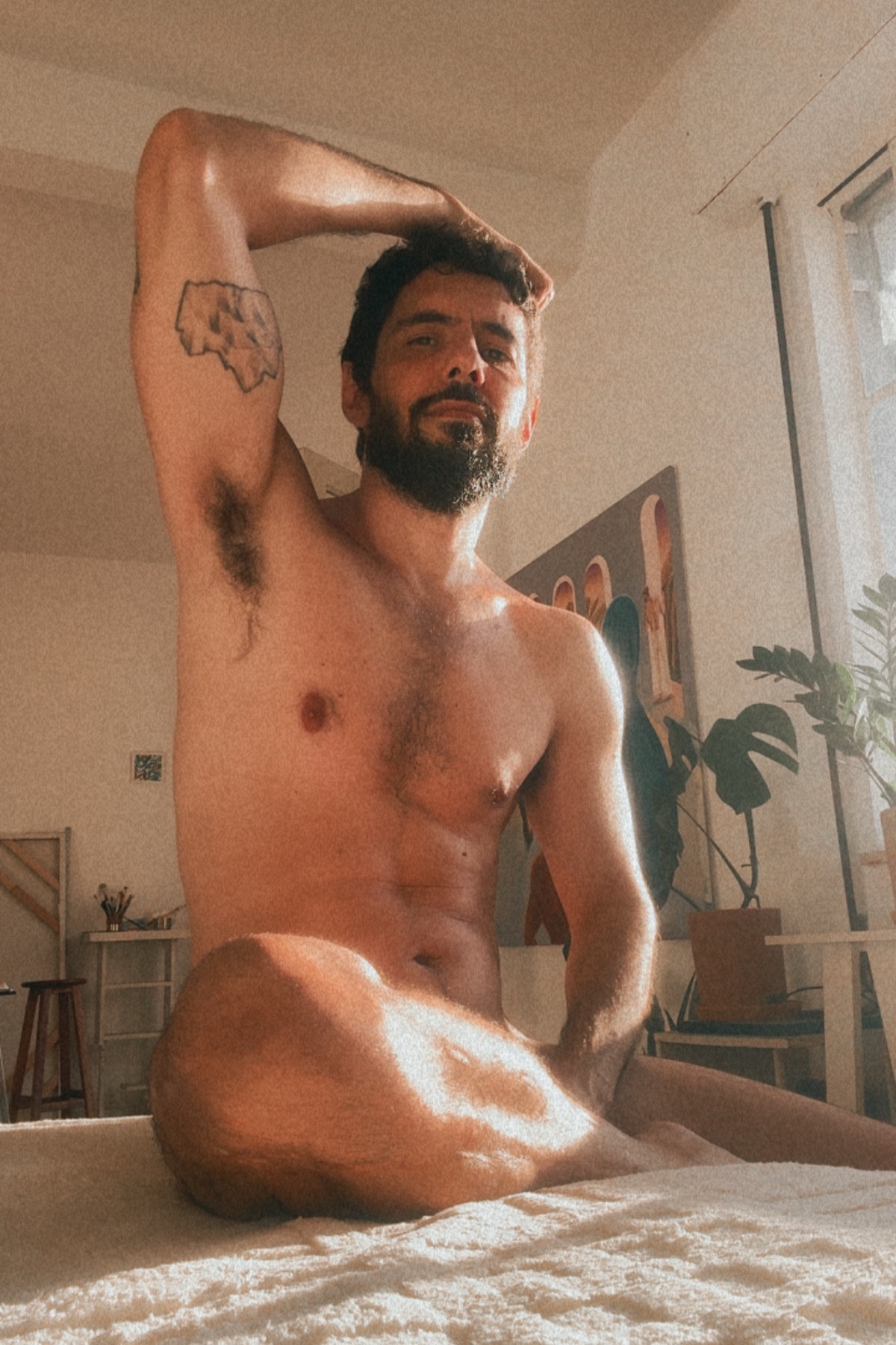 A man with a beard and a tattoo on his arm poses indoors, bathed in natural light.