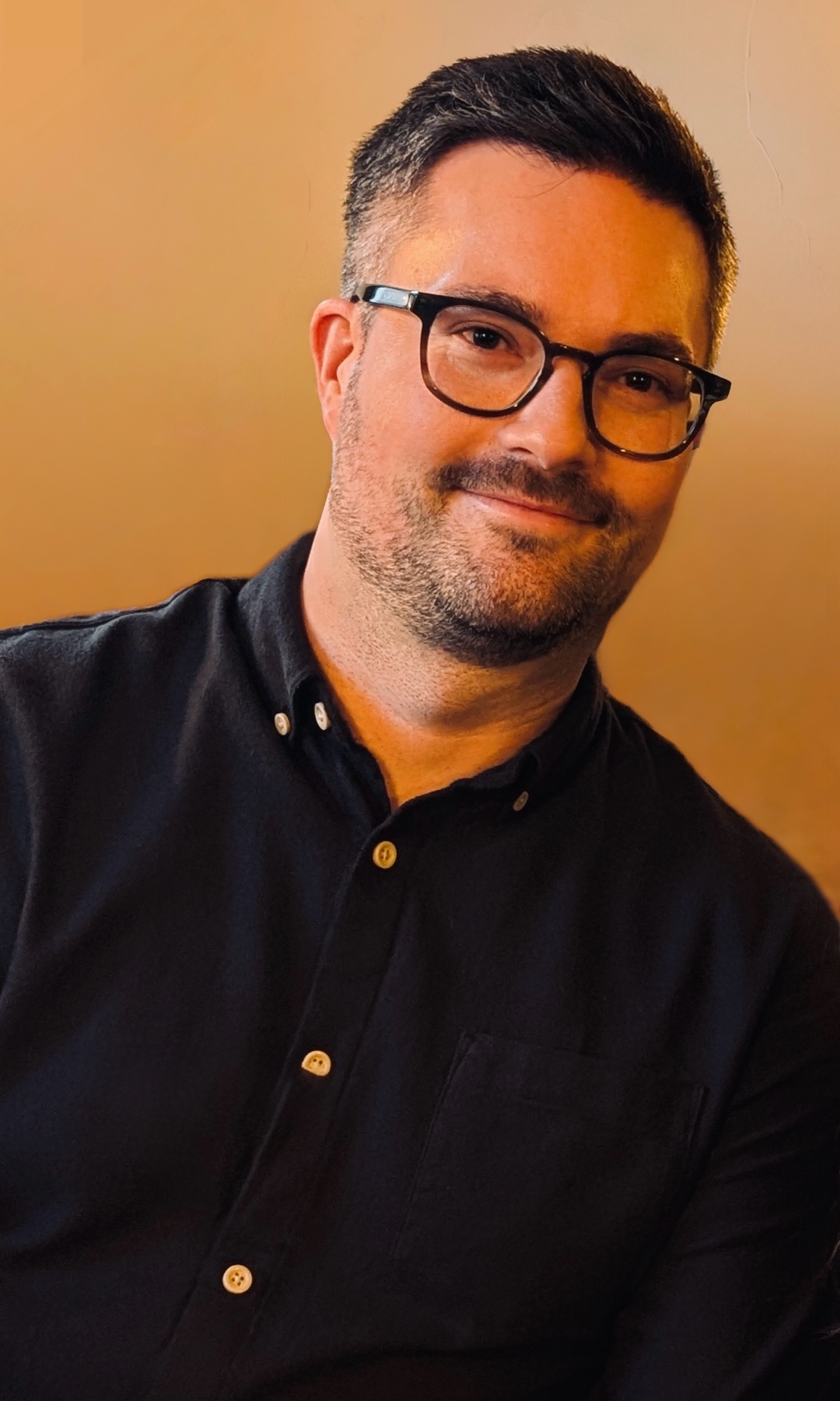 A portrait of a man wearing glasses and a black shirt. He is looking at the camera.