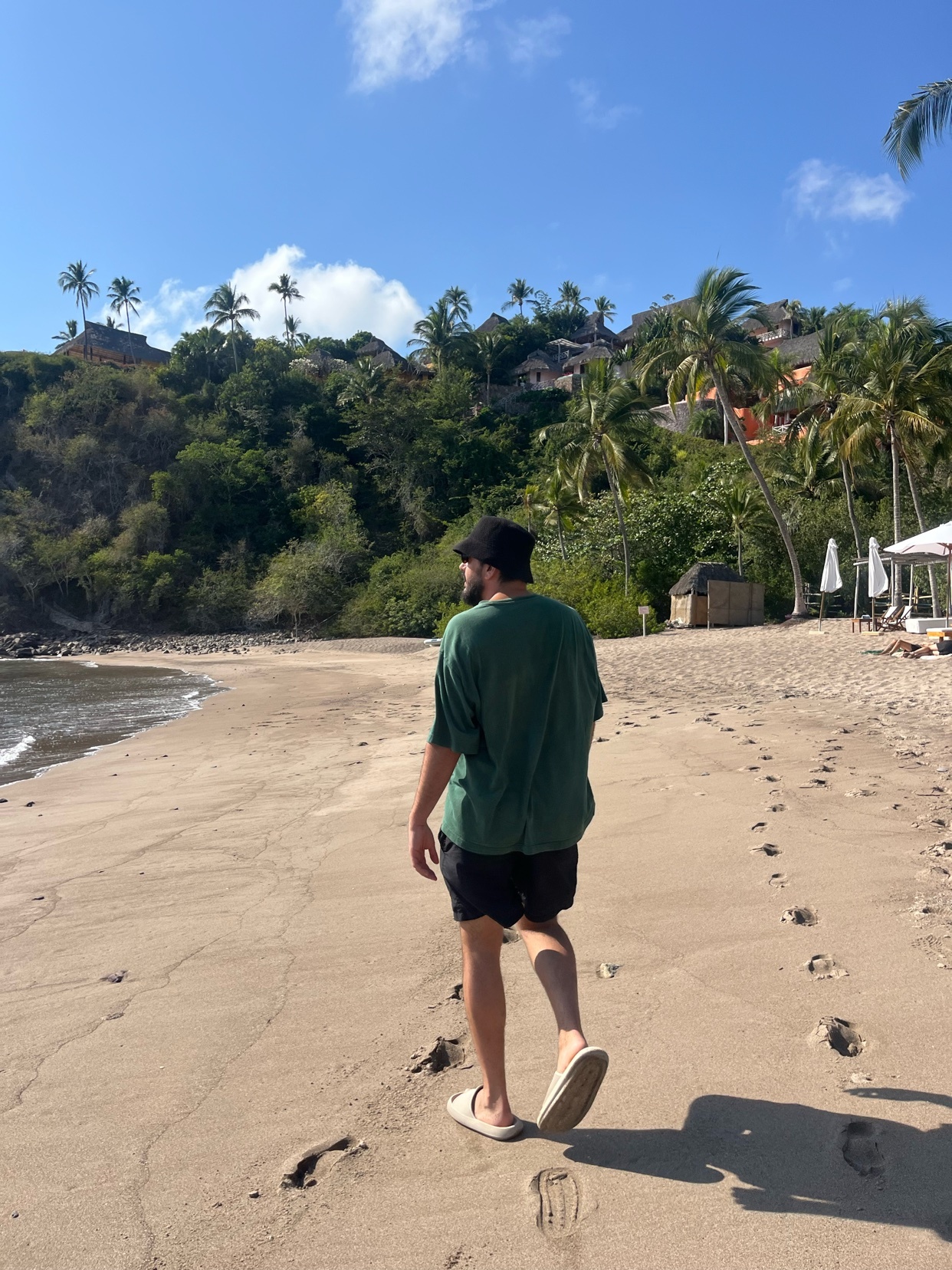 A man walks along a sandy beach, leaving footprints behind him. Lush green hills dotted with buildings and palm trees rise behind the beach under a clear blue sky.
