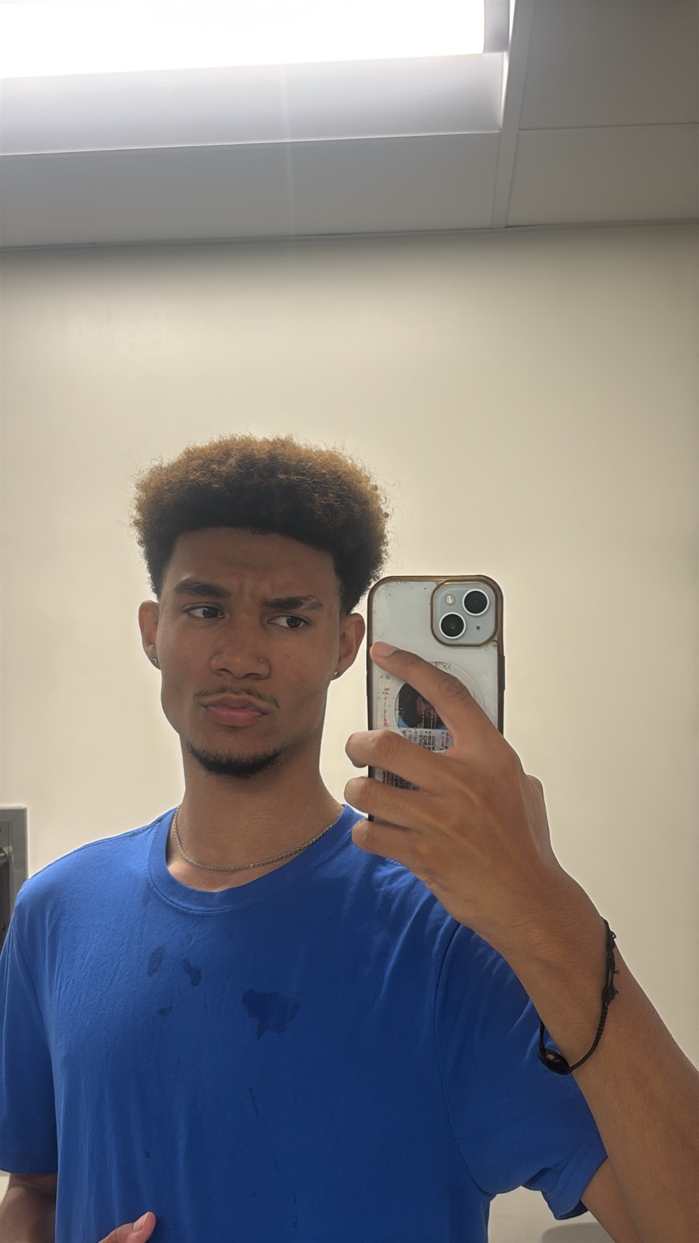A young man with curly hair and stubble takes a selfie in a bathroom, wearing a blue t-shirt.