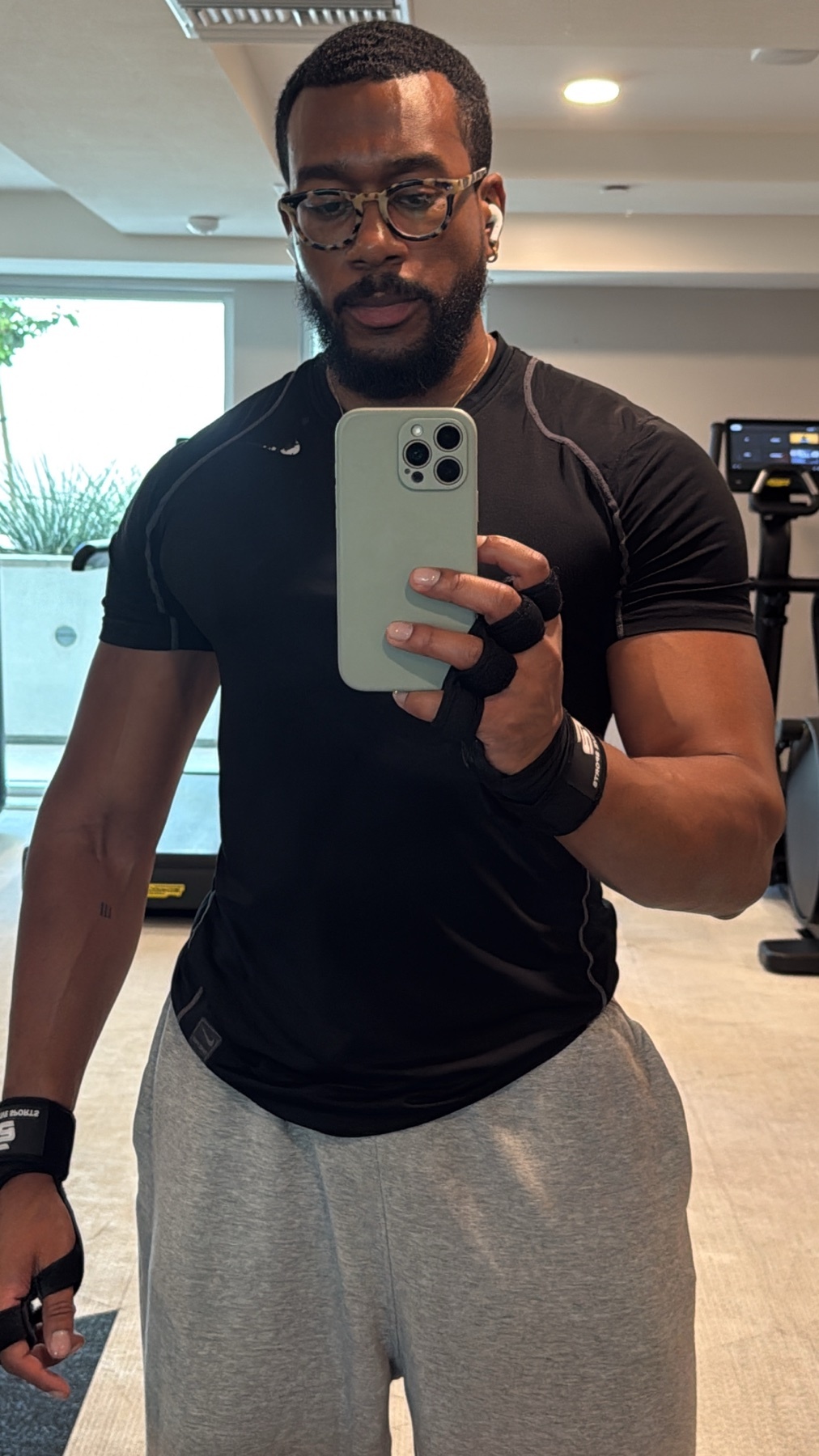 A man with a beard and glasses takes a selfie in a gym, wearing a black t-shirt and grey sweatpants.