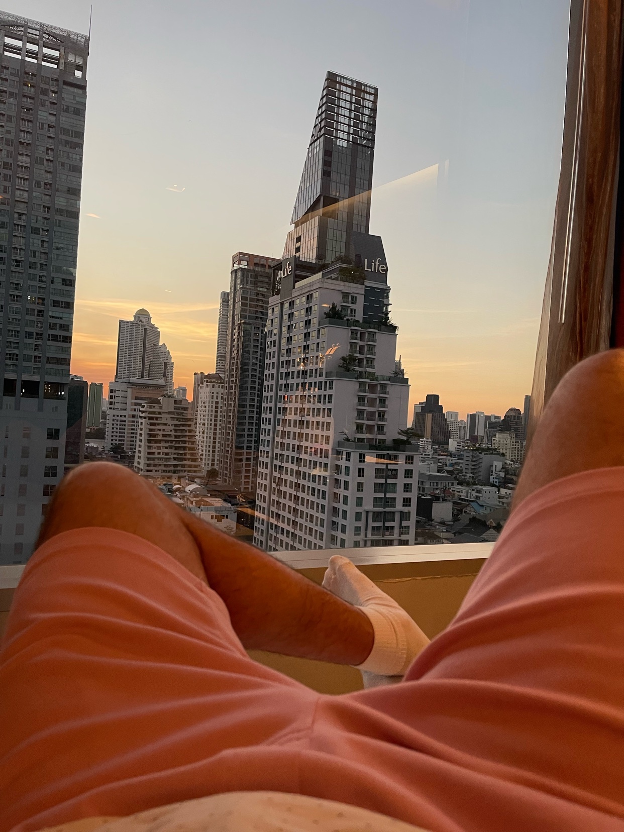 A man relaxes in a hotel room, with a cityscape visible through the window. The photo highlights the man's legs and shorts, with a suggestive focus.