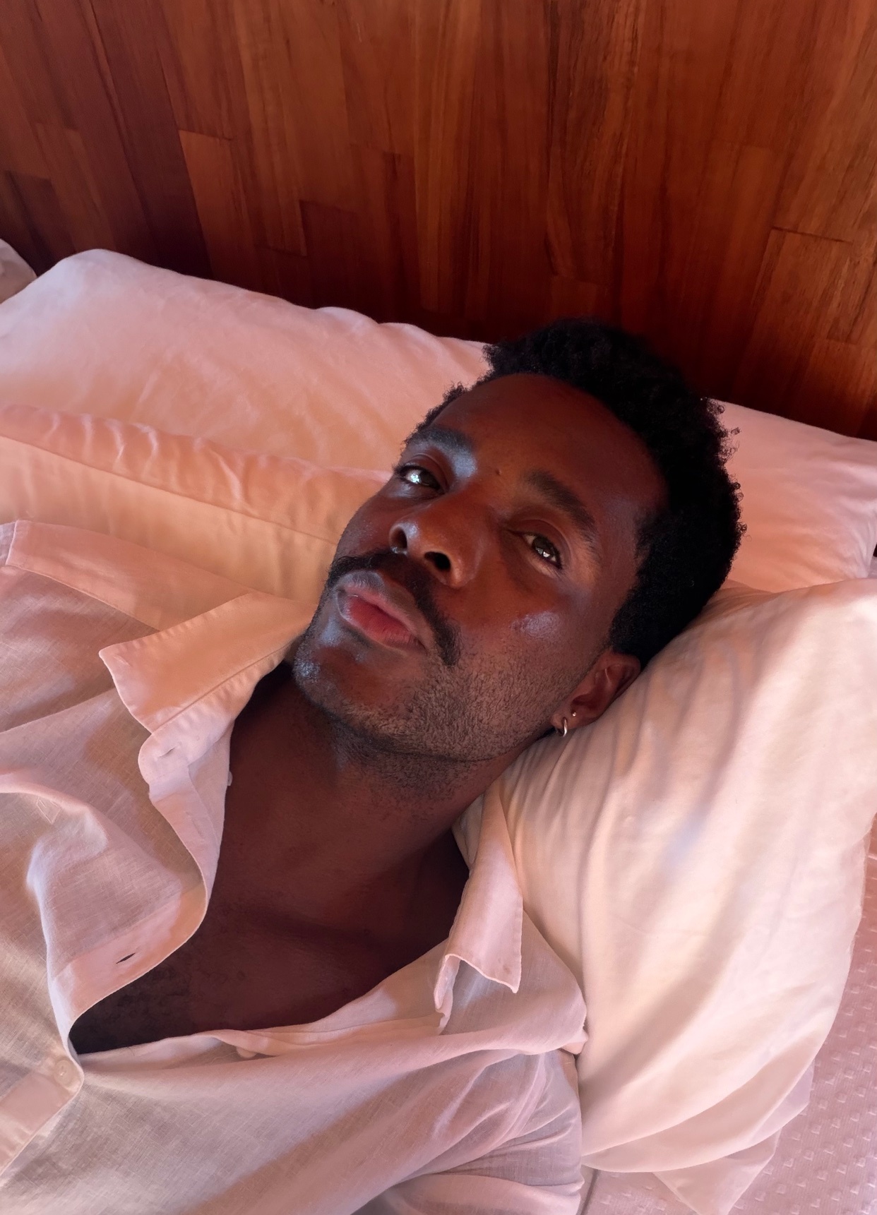 A man with dark hair and stubble lies on a pillow in bed, looking up with his mouth slightly open. He is wearing an unbuttoned white shirt.