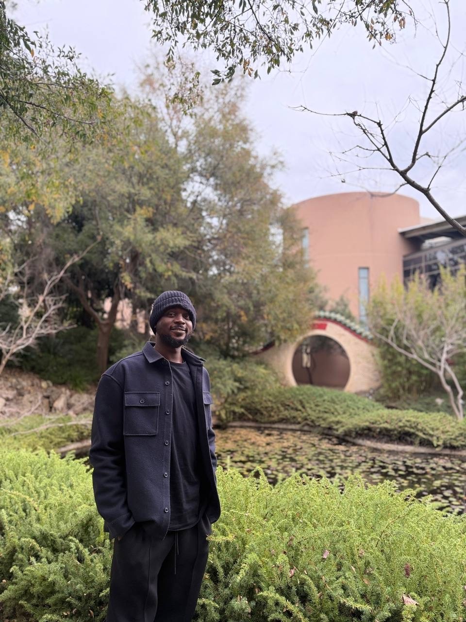 A Black man wearing a black beanie and black clothing stands in a lush garden with a pond and a unique building in the background.