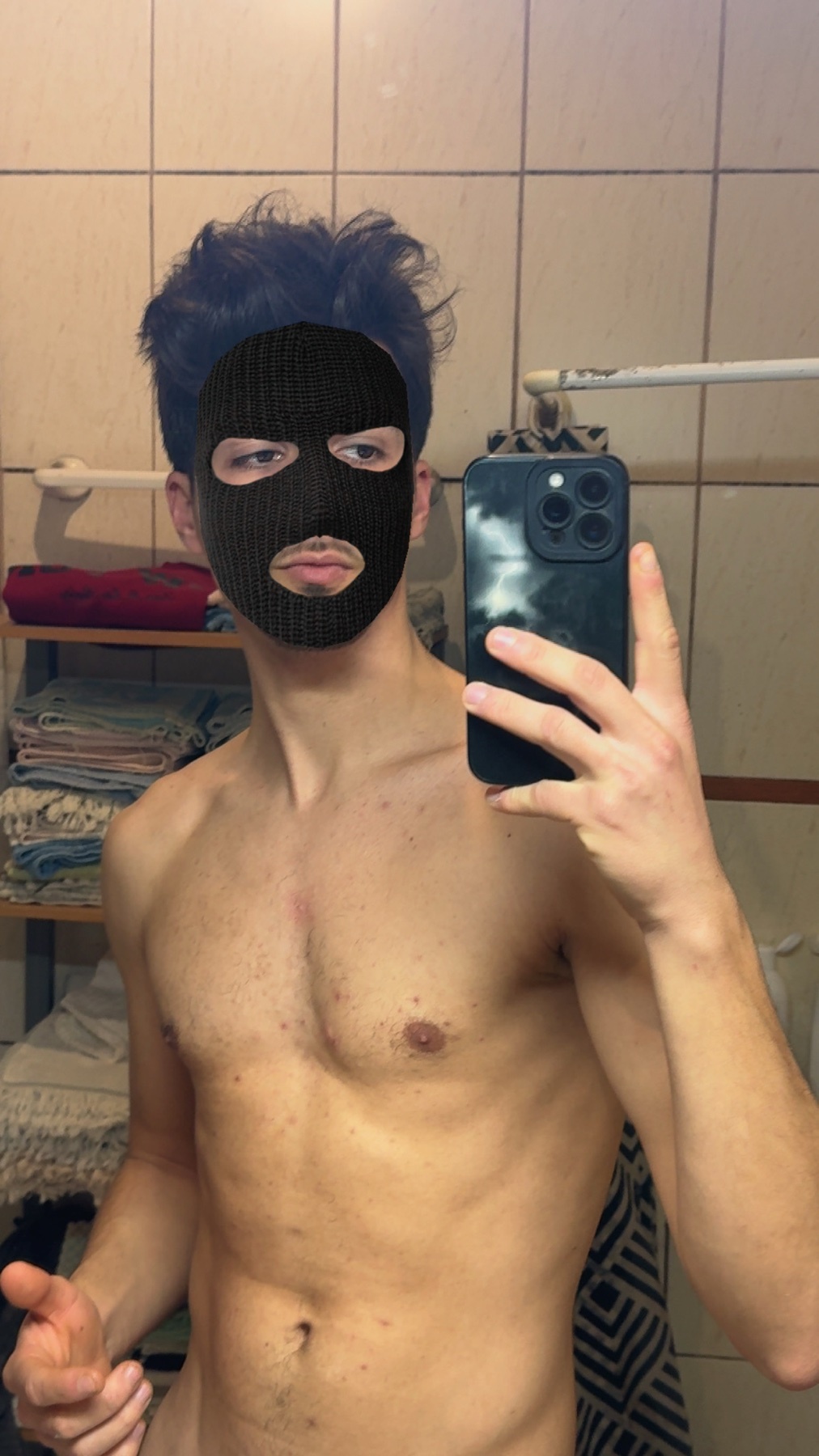 A shirtless man takes a selfie in a bathroom, wearing a black balaclava. He is flexing his arm.