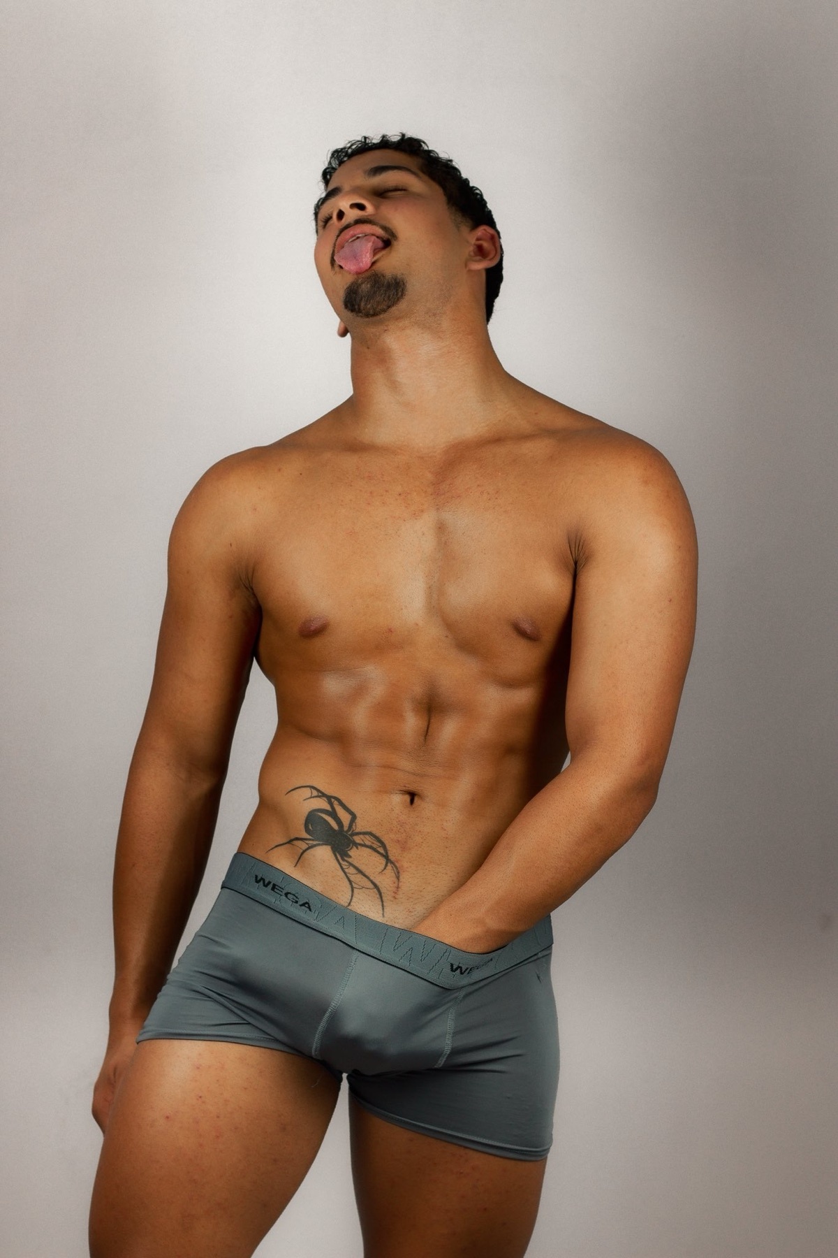 A man with a spider tattoo on his abdomen poses in grey boxer briefs, sticking his tongue out with his eyes closed.