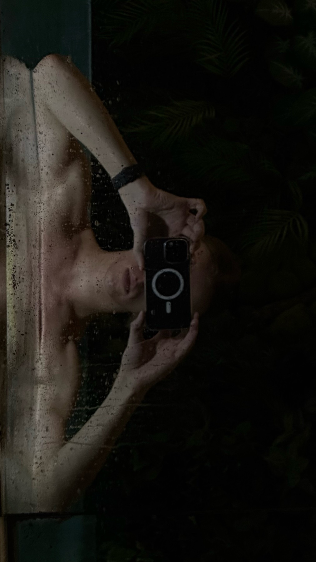 A shirtless man takes a selfie in front of a mirror. The photo is dark, with water droplets on the mirror and a tropical background.